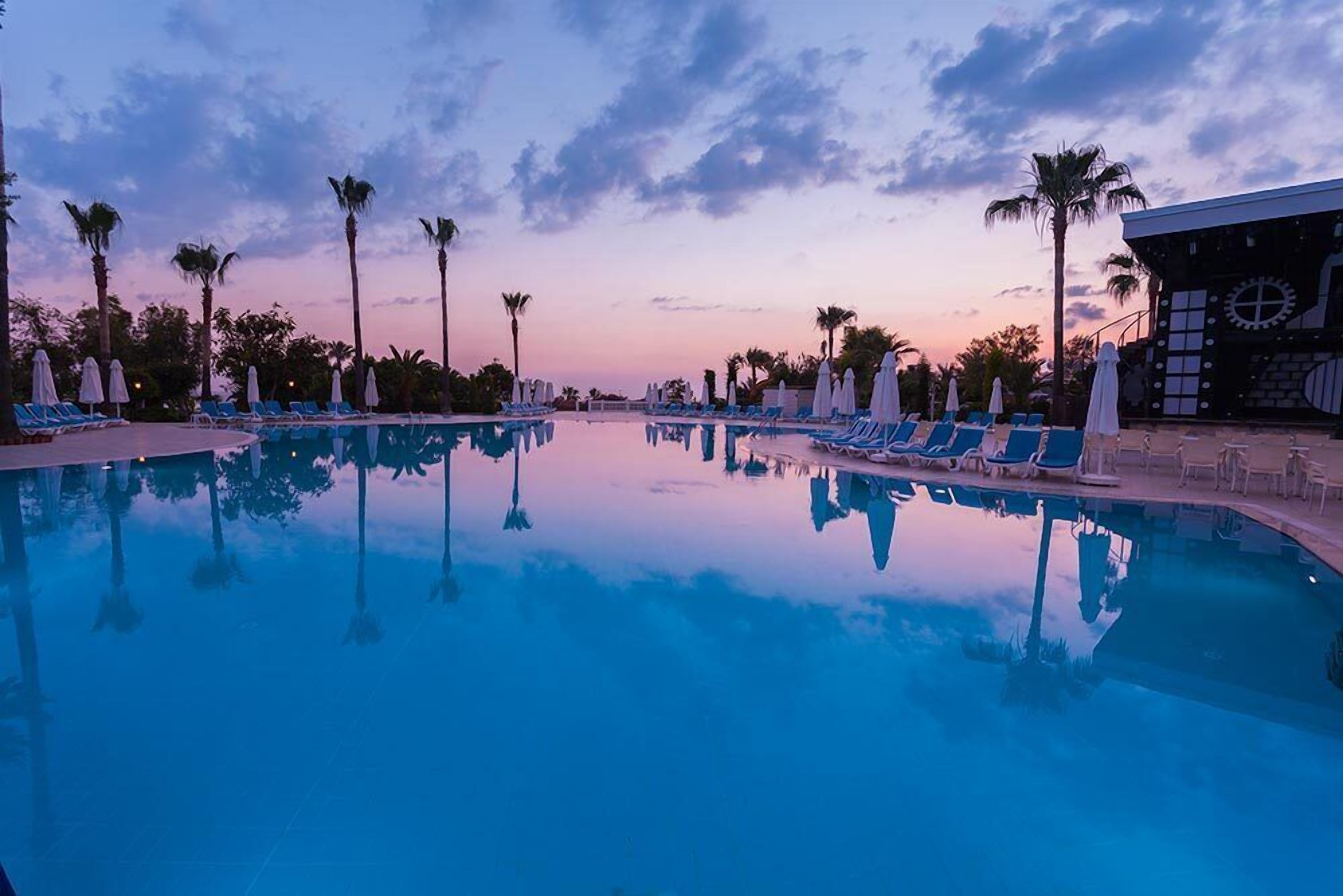 Zona de piscina con sillas de descanso y sombrillas, rodeada de palmeras que reflejan el cielo.