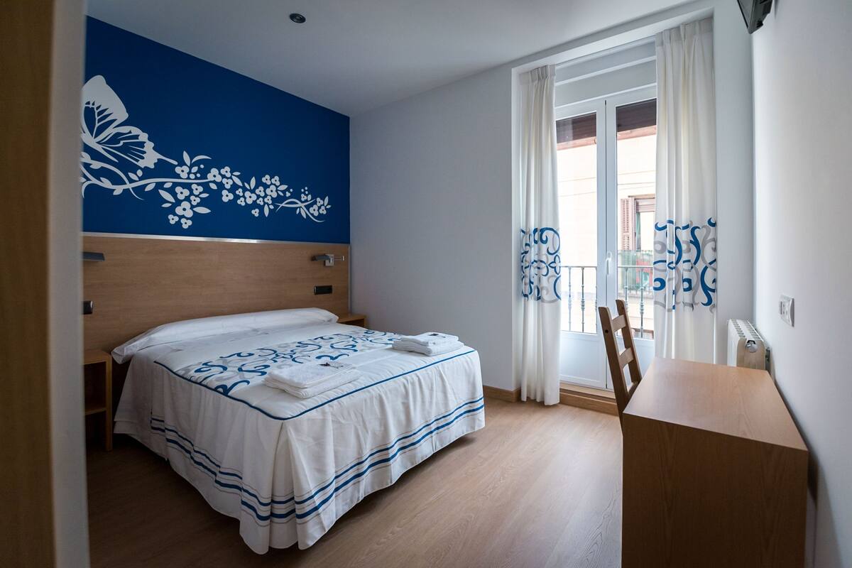 A hotel room with a bed, a wooden desk, a window with curtains, and a wall with a floral design.