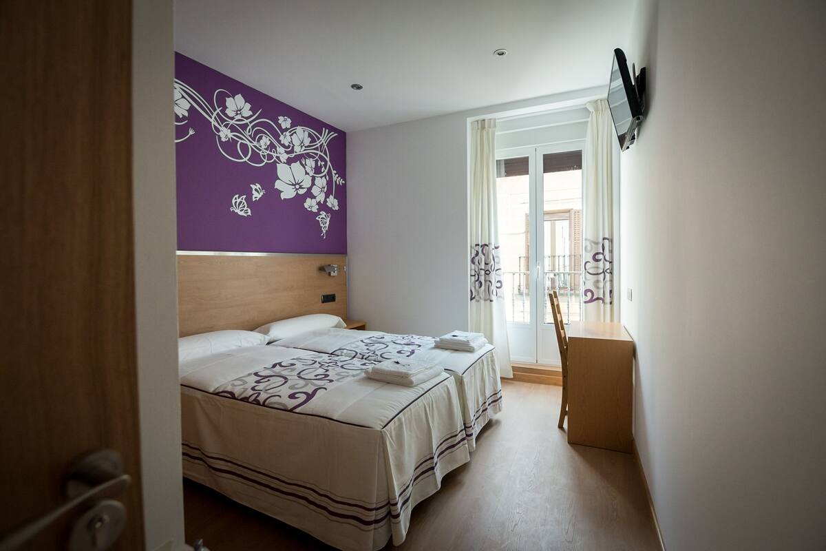 A hotel room with two beds, a wooden headboard, a purple floral wall, a window with curtains, and a wooden chair.