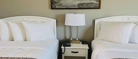Habitación de hotel con dos camas, una mesita de noche con lámpara, un teléfono y un cuadro de una escena portuaria.