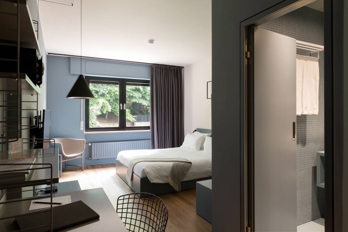 Een moderne hotelkamer met een bed, een bureau met stoel, een raam met gordijnen en uitzicht op groen.