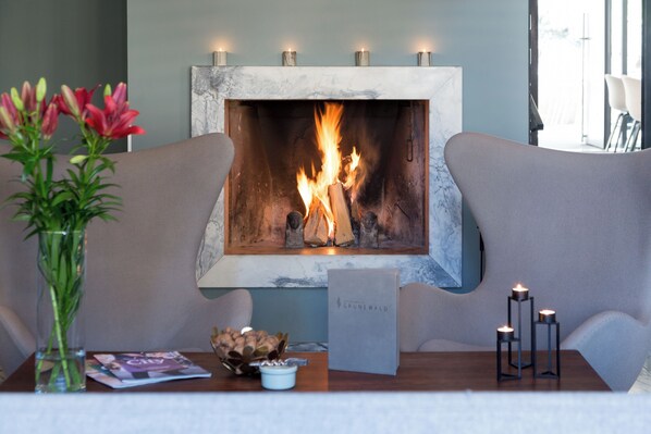 A fireplace with a fire, a vase of flowers, a magazine, and a Grunewald sign on a wooden table. - Hostellerie du Grünewald (Luxembourg City)