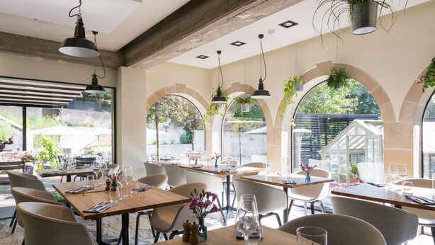 Un restaurante con mesas y sillas de madera, plantas colgantes y ventanas grandes con marcos arqueados.
