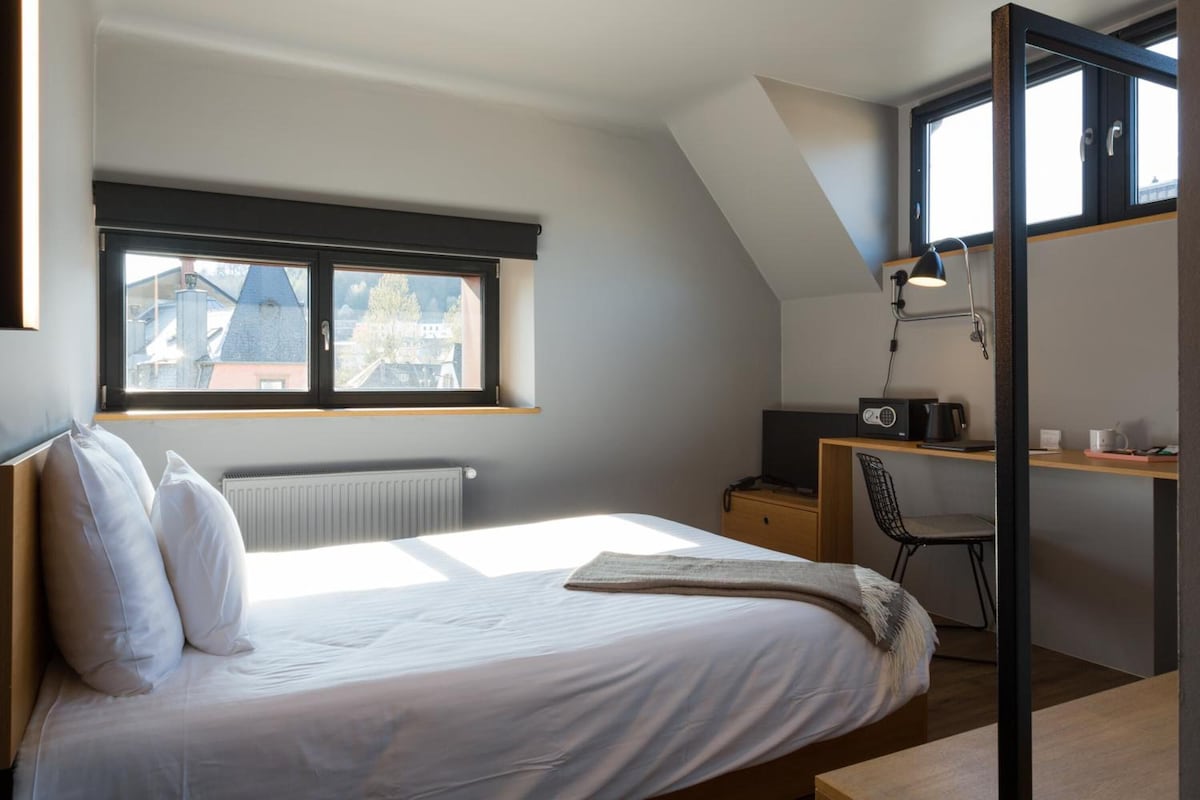 A bedroom with a bed, a desk, a chair, a radiator, and a window with a view of buildings.