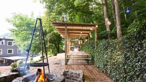 Un patio al aire libre con un hogar de piedra, una pérgola de madera y una mesa dispuesta para comer, rodeado de follaje.