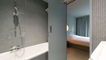 A modern bathroom with a shower, a bathtub, and a visible bed in the adjacent room.