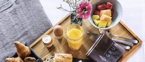 Uma bandeja de café da manhã com croissants, uma tigela de cereal, um copo de suco de laranja, um pequeno vaso com flores e um prato de frutas.