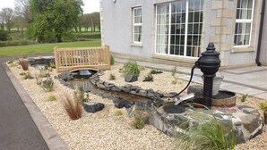 A landscaped garden with a pond, a wooden bench, and a stone pathway leading to a building with large windows. - Ballyharvey House B&B (Antrim)