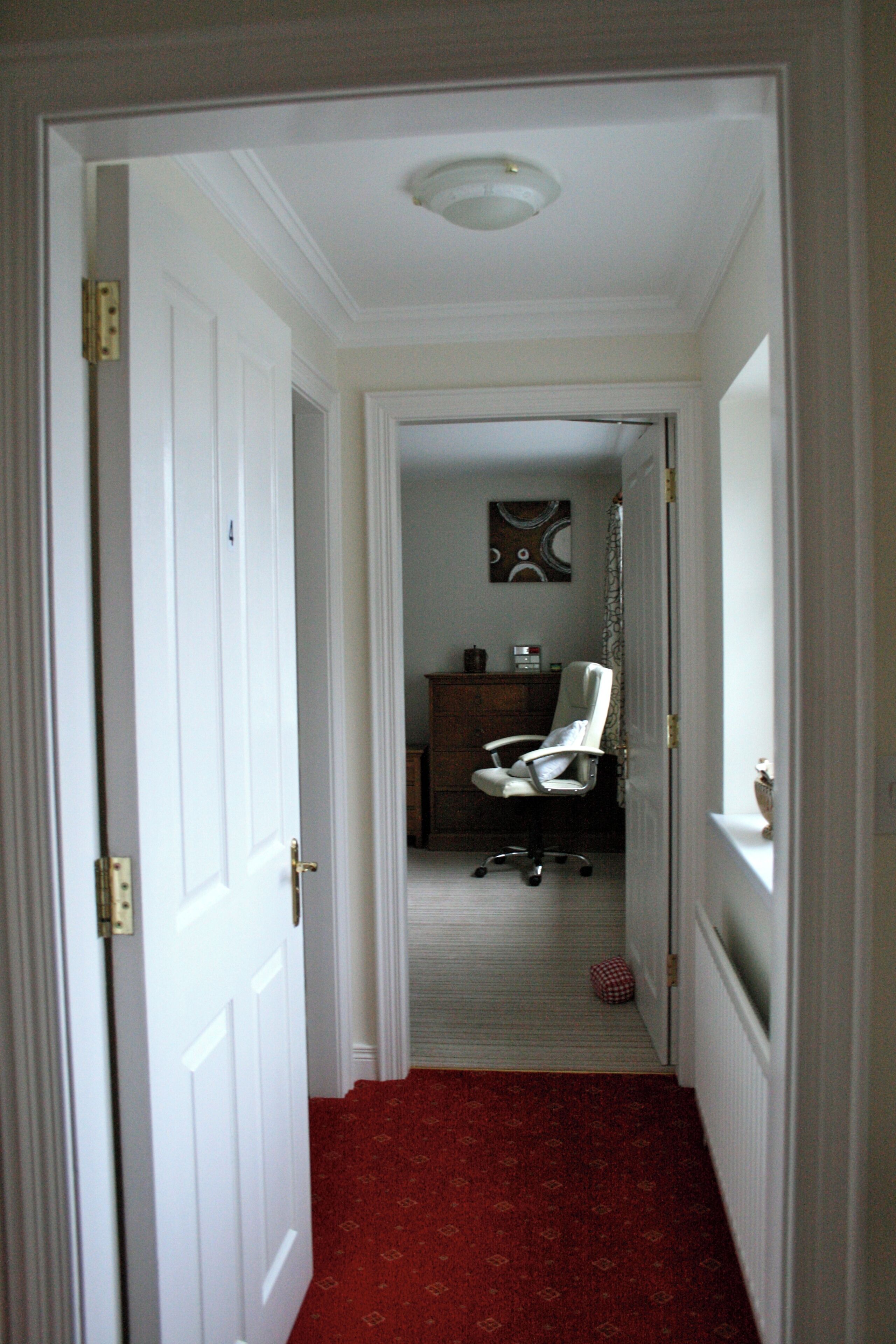 A hallway with a red carpet leading to a room with a white chair and a dresser.