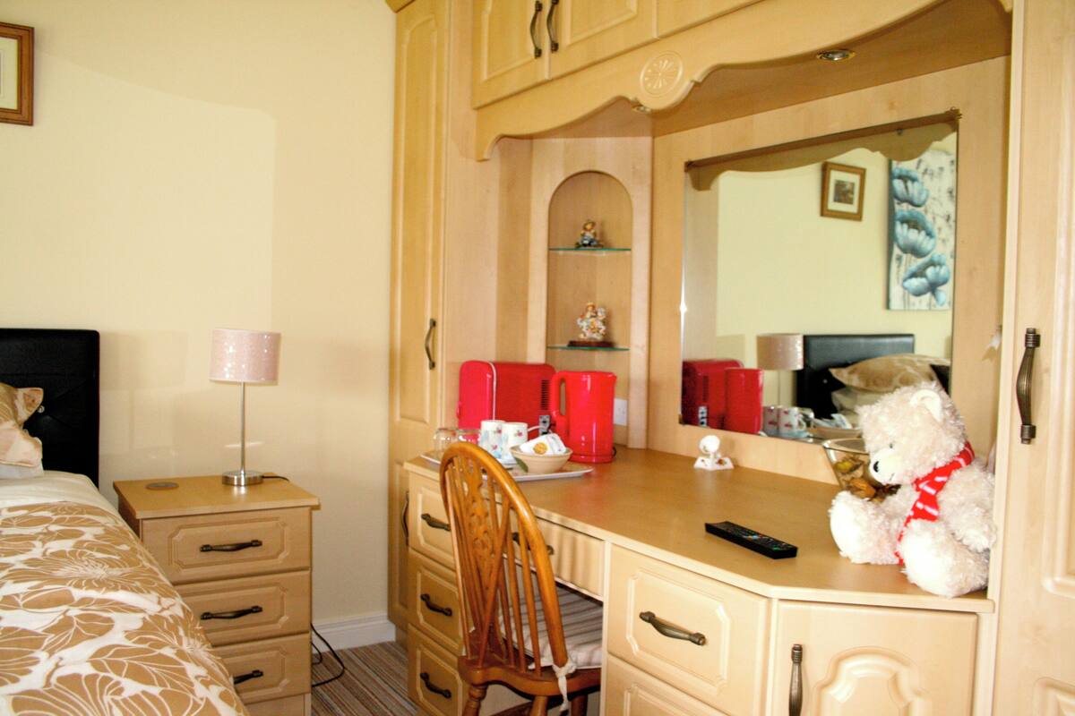 Une chambre d’hôtel avec un lit, une commode en bois, un miroir, une chaise, un ours en peluche et une machine à café rouge.