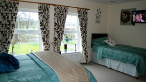 A bedroom with two beds, a window with curtains, a television on the wall, and a vase with flowers on the windowsill. - Ballyharvey House B&B (Antrim)