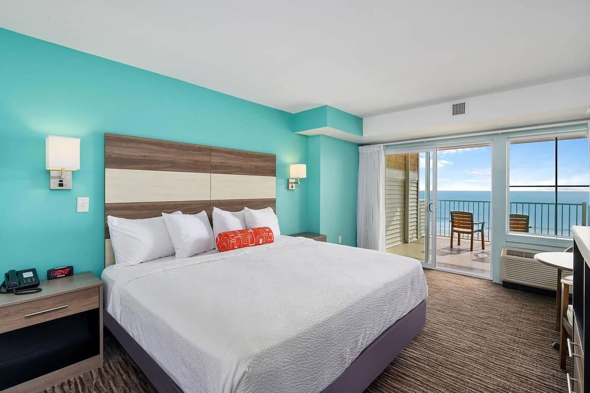 Une chambre d’hôtel avec un grand lit, une table de chevet avec un téléphone, une horloge numérique et une vue sur l’océan par une porte coulissante.