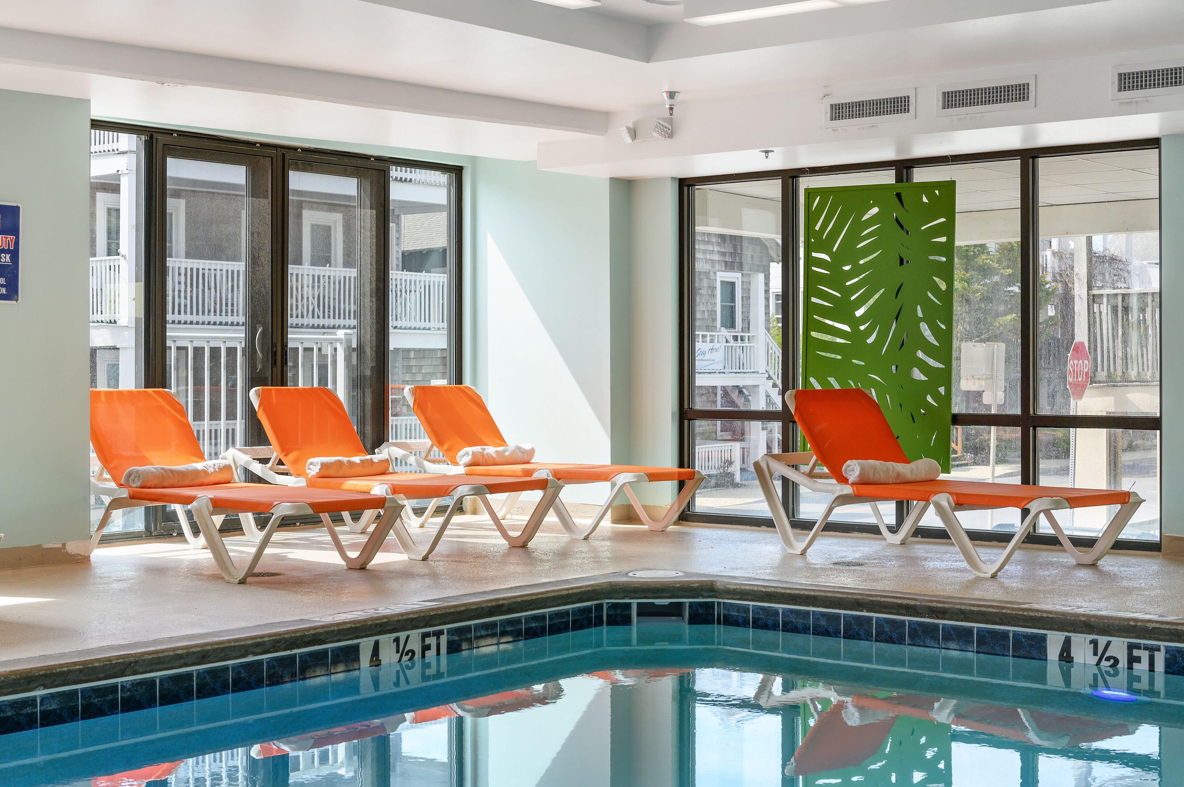 Zona de piscina con cuatro sillones reclinables color naranja y un panel decorativo verde.
