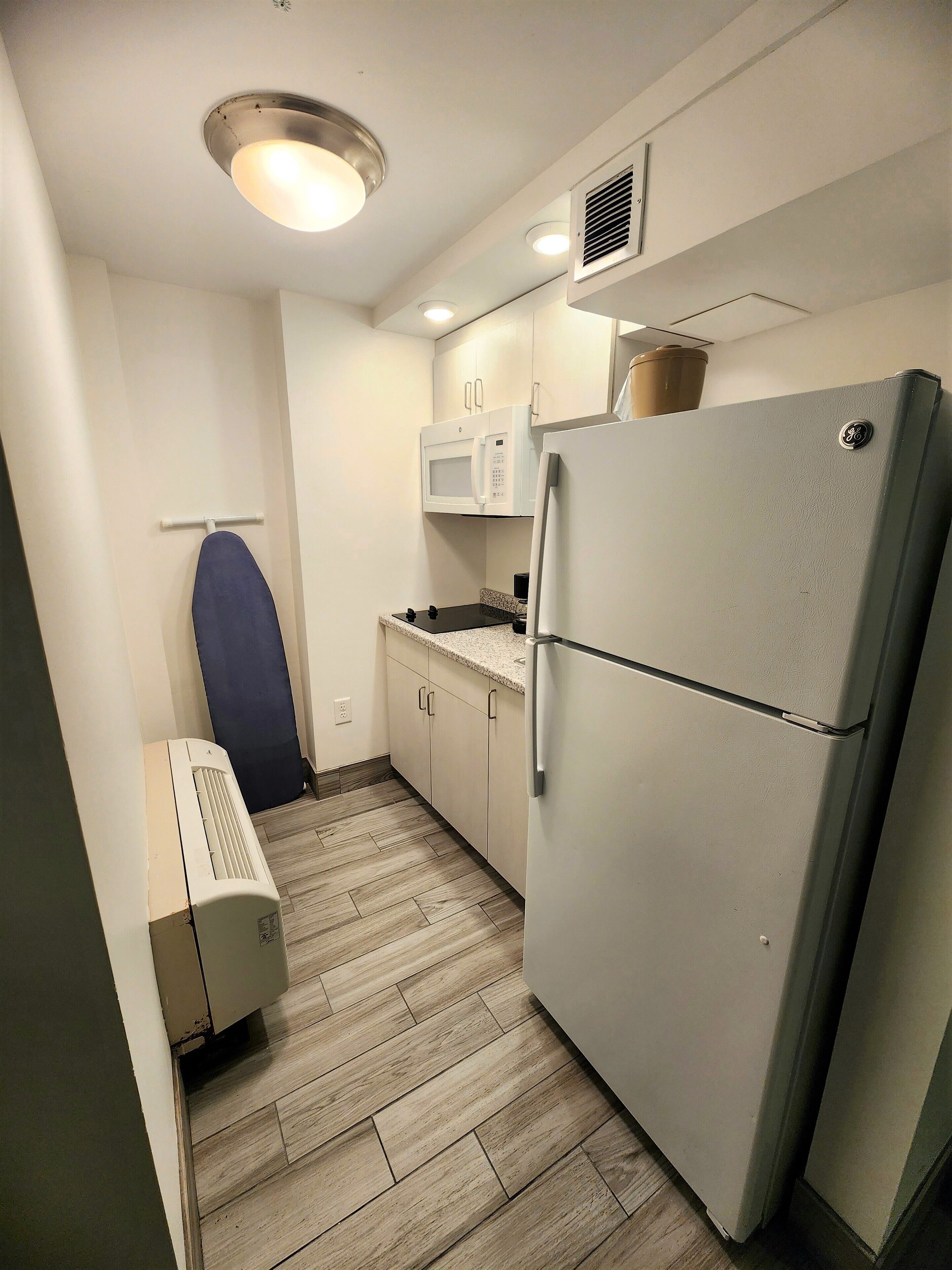 A compact kitchen with a microwave, refrigerator, and ironing board.