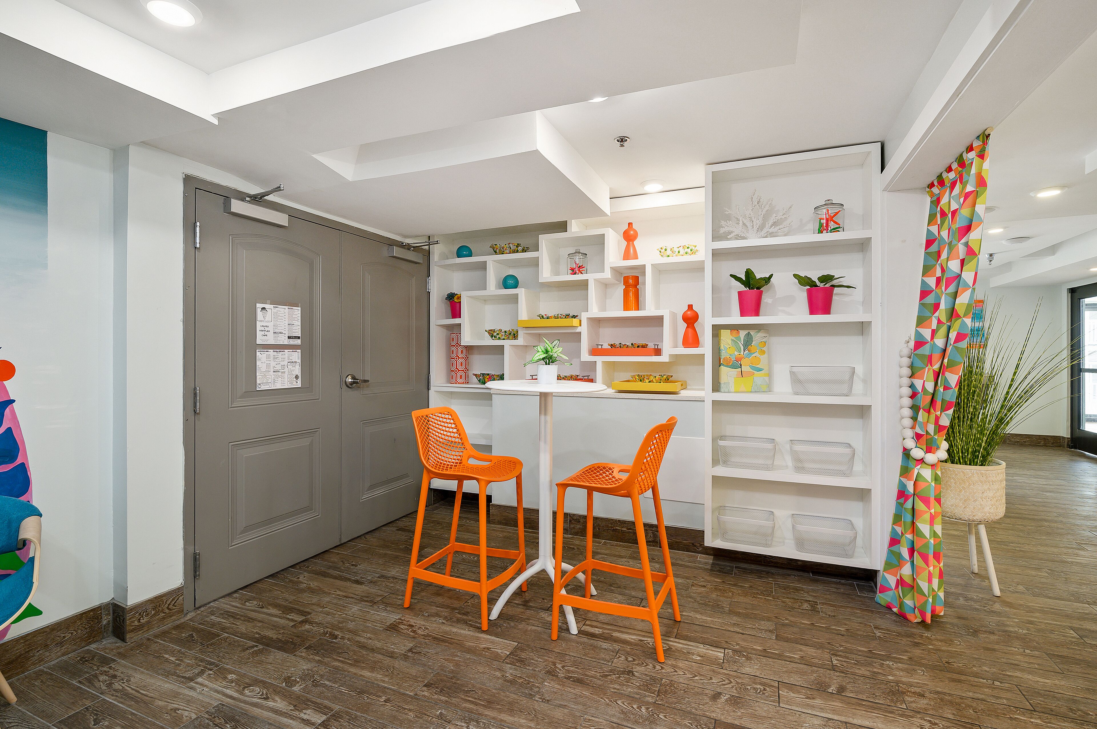 A modern interior with a grey door, two orange chairs, a white table, and a colorful plant.