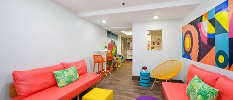 A modern lounge area with red sofas, a yellow ottoman, and a purple stool. A colorful abstract wall art is visible.