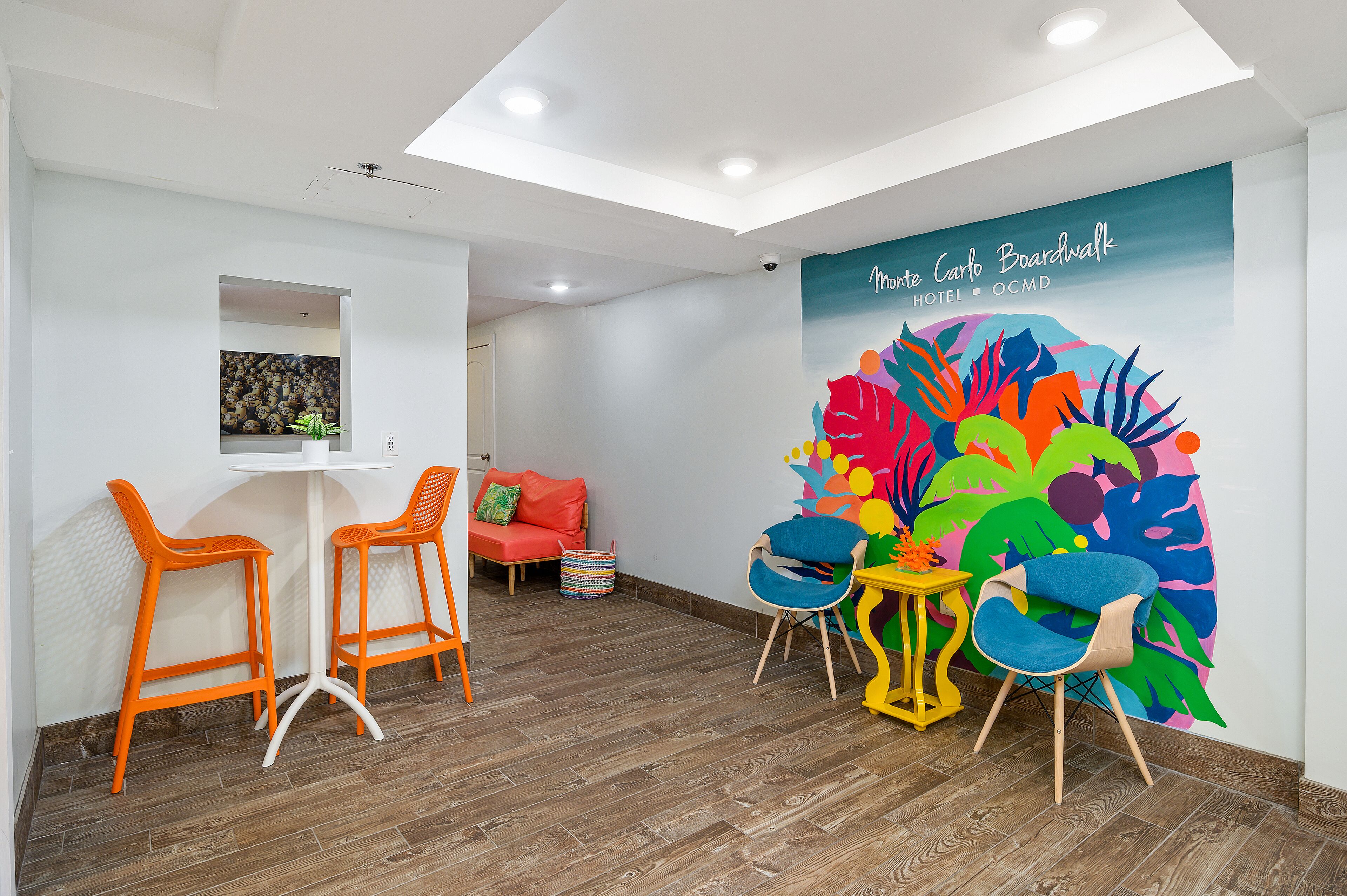 A modern hotel room with a colorful mural, wooden flooring, and a mix of orange and blue chairs.