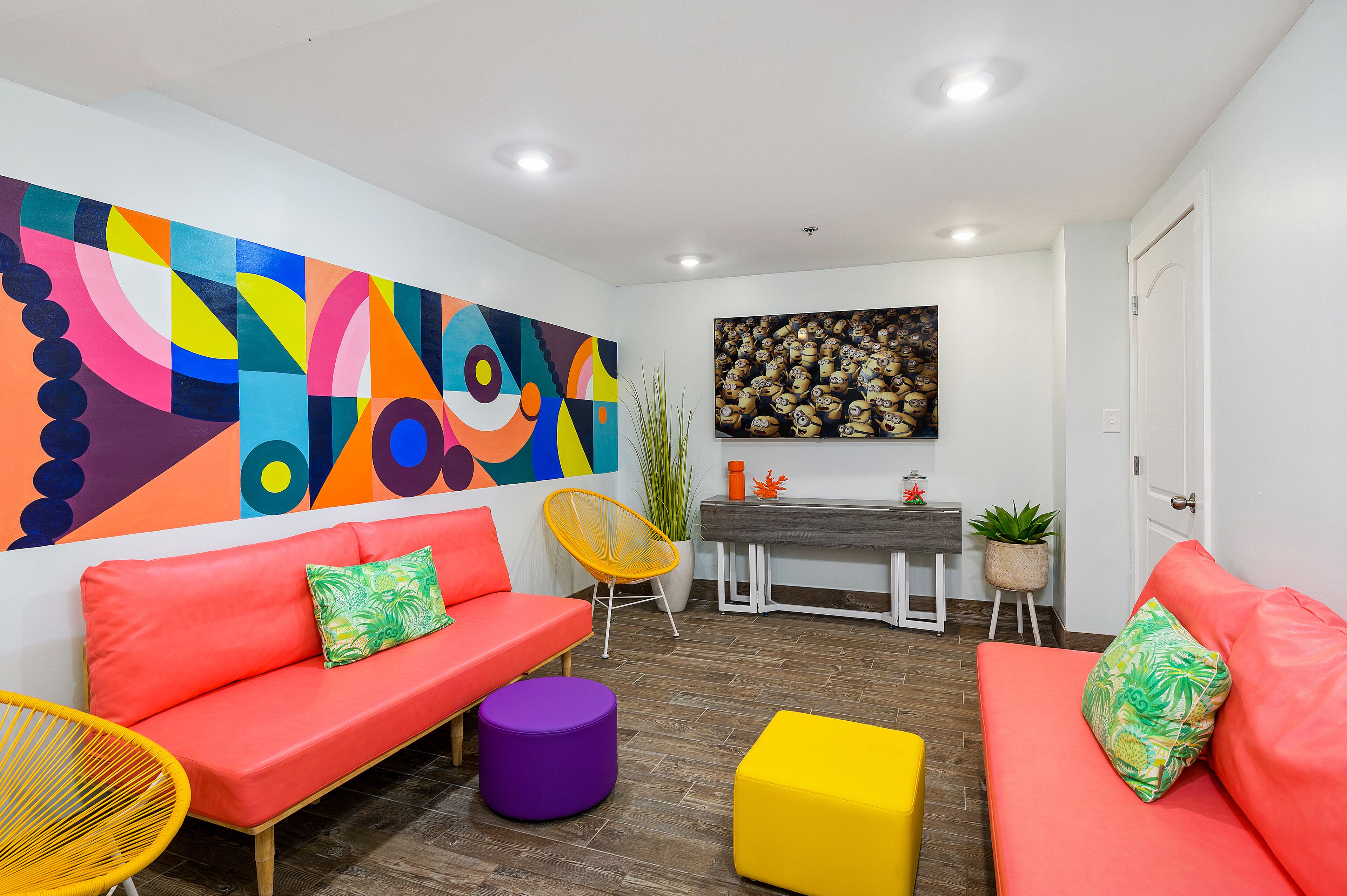 Un salon moderne agrémenté d’une fresque murale abstraite aux couleurs vives, d’une table à manger en bois et d’un coin salon confortable avec un canapé rouge et des coussins colorés.