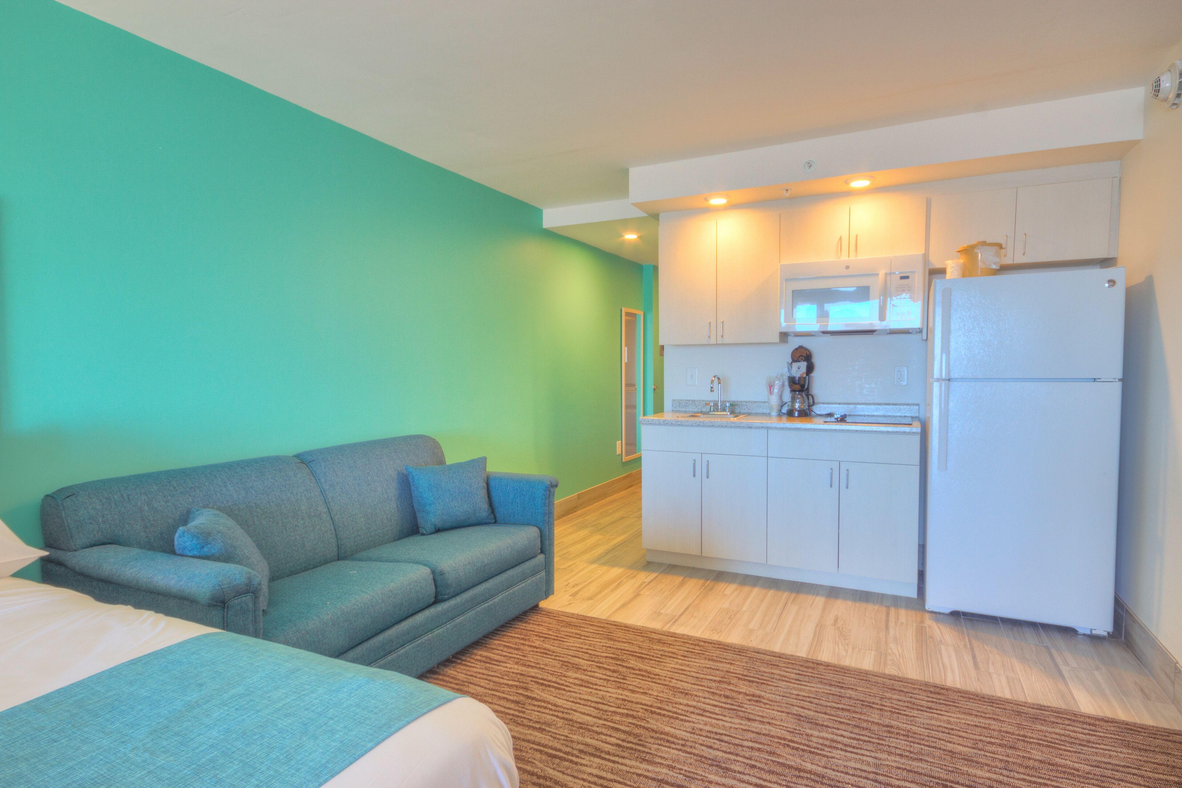 Habitación compacta con sofá, cama, kitchenette con refrigerador, microondas y armarios, y piso de madera.