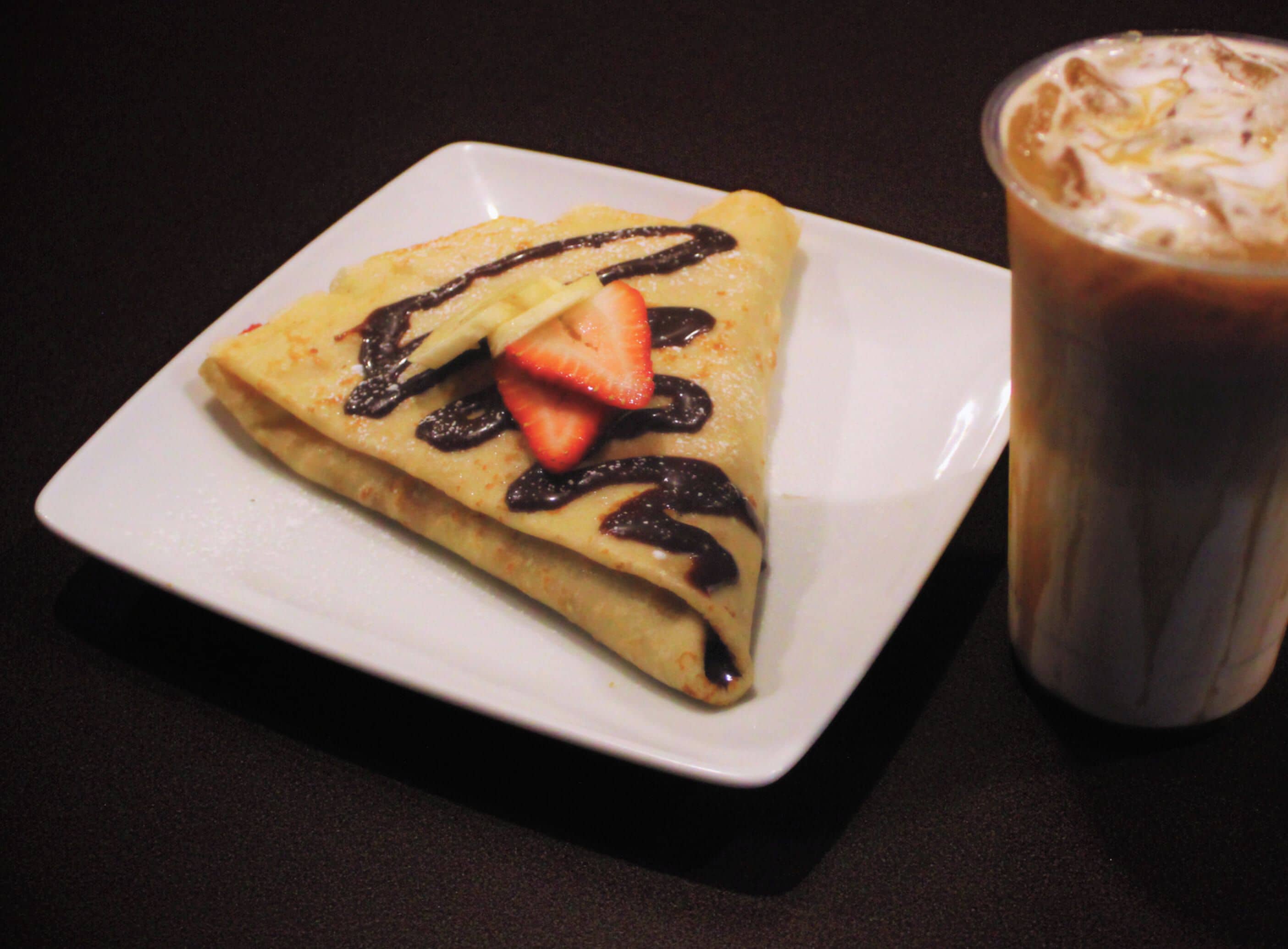 A square plate with a folded crepe topped with chocolate and a strawberry, and a glass of iced coffee beside it.