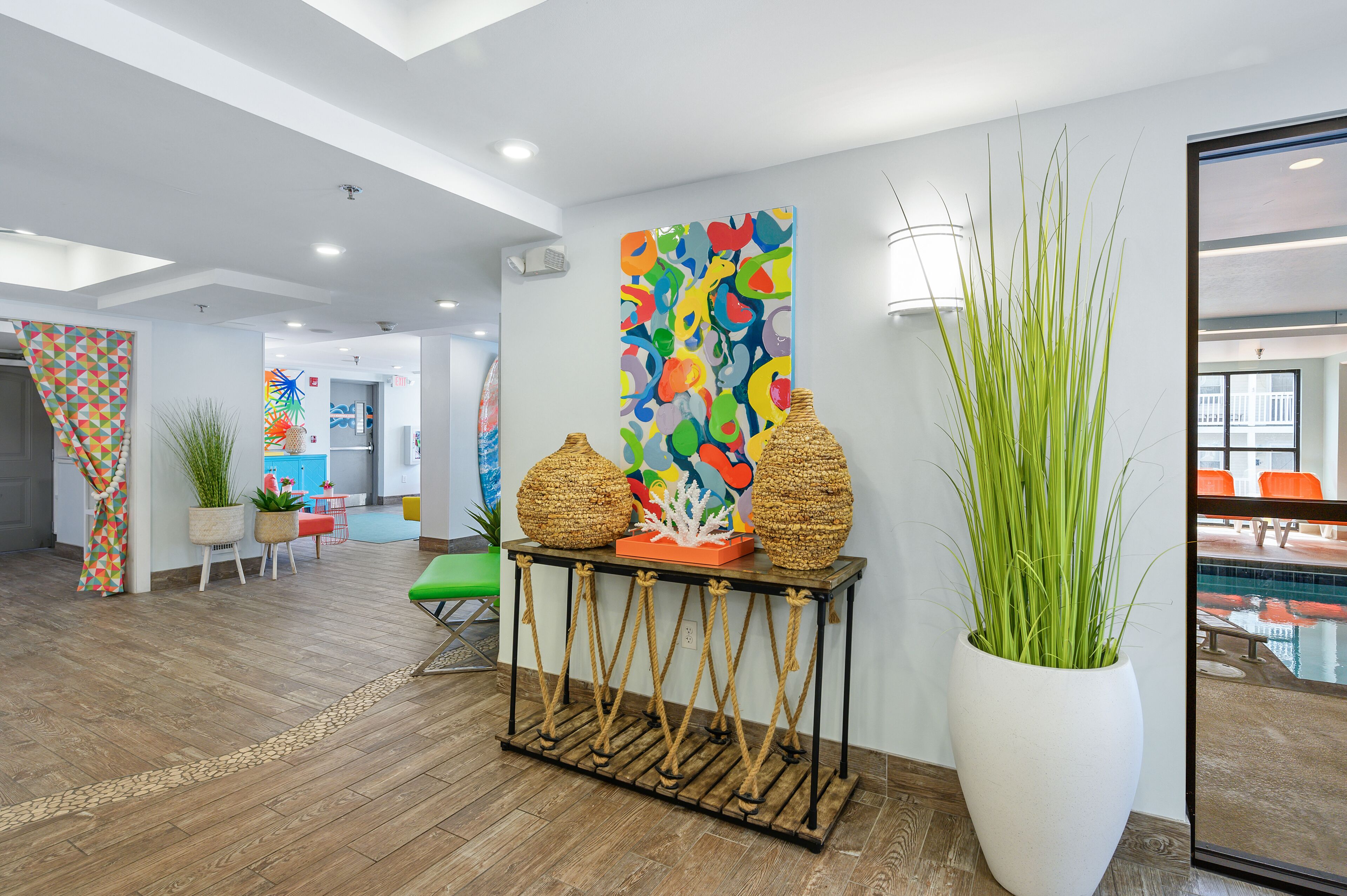 Um lobby moderno com piso de madeira, um vaso branco grande com plantas verdes, uma pintura abstrata colorida e um espelho grande refletindo uma área de piscina.