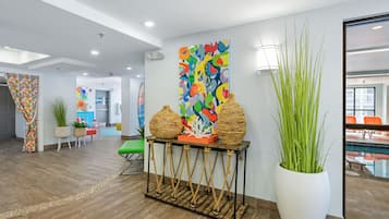 Un hall moderno con suelo de madera, un gran jarro blanco con plantas verdes, un cuadro abstracto colorido y un espejo grande que refleja una zona de piscina.