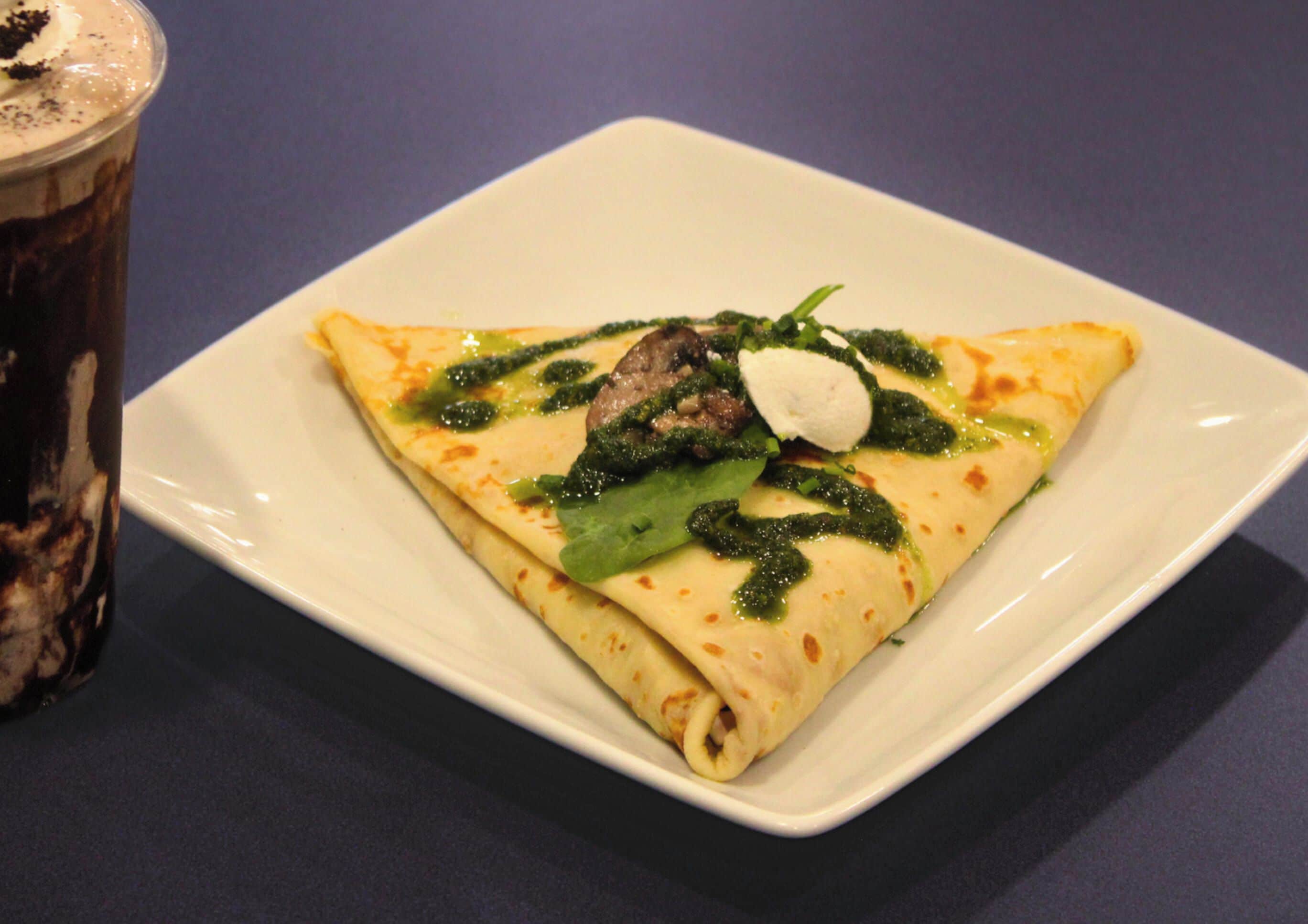 Une assiette carrée blanche sur laquelle repose une crêpe pliée, garnie de légumes verts, de champignons et d’une cuillerée de crème, accompagnée d’un verre d’une boisson sombre.