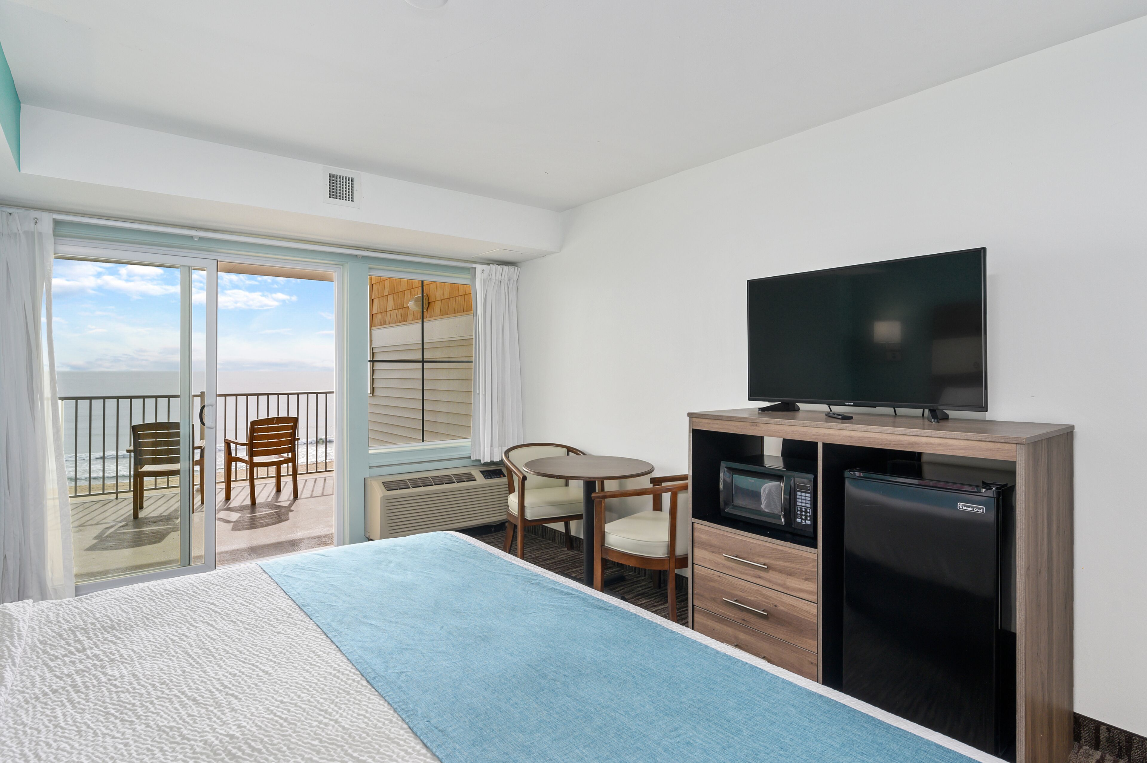 Habitación de hotel con cama, televisión, refrigerador, microondas, mesa con sillas y balcón con vista al océano.