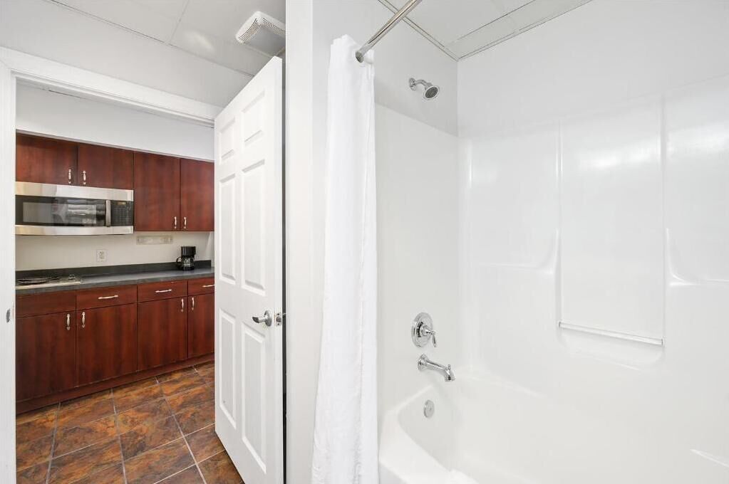 A bathroom with a bathtub and shower, a white door, and a kitchen area with wooden cabinets and a microwave.