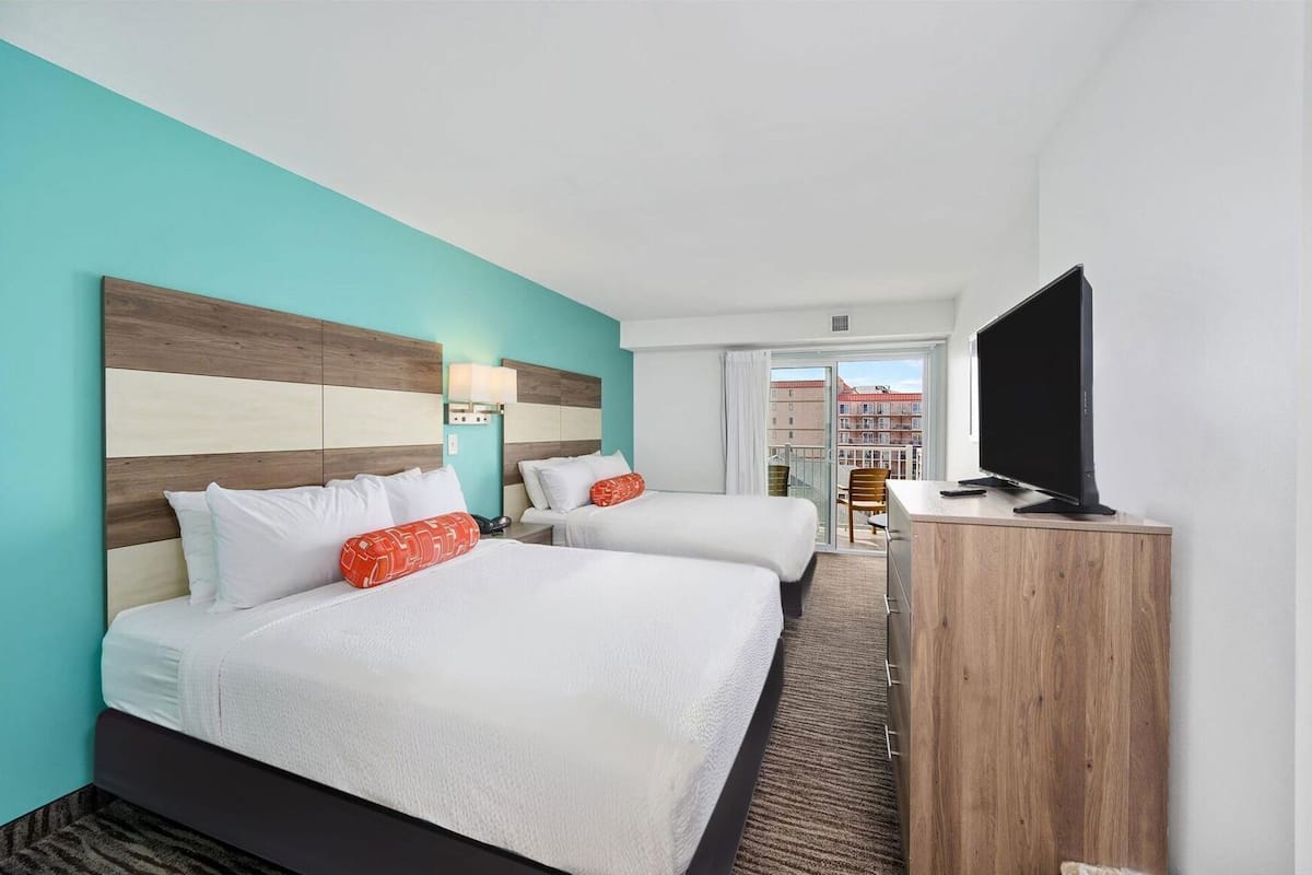 Une chambre d’hôtel avec deux lits, un téléviseur posé sur un meuble en bois, et une vue sur les immeubles à travers une porte vitrée.