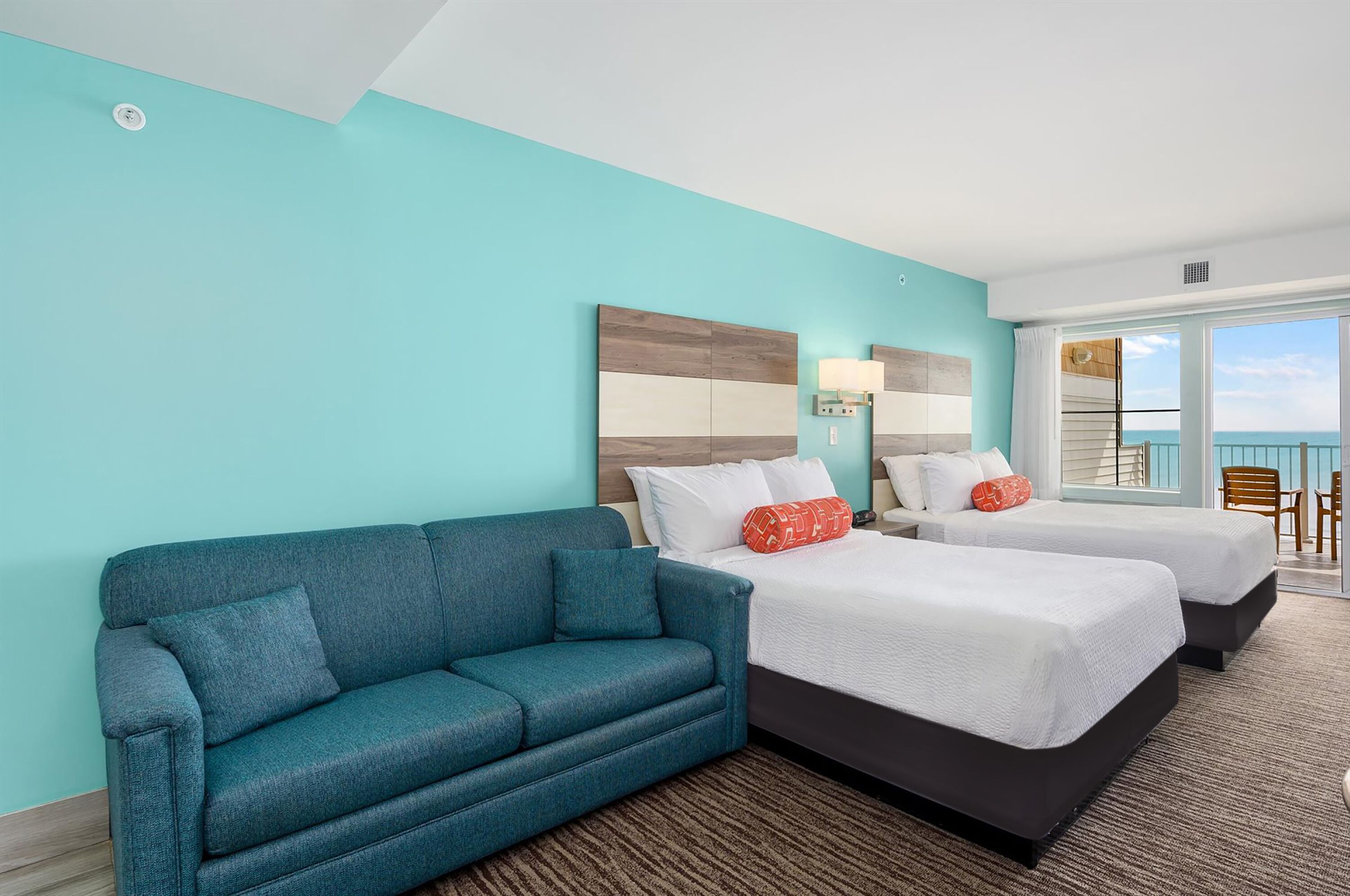 A hotel room with a teal wall, two beds, a blue sofa, and a large window offering a view.