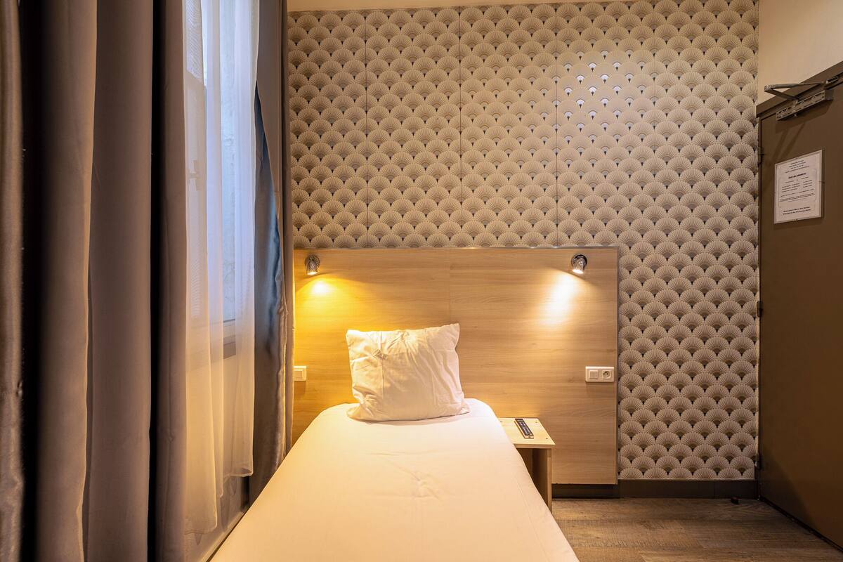 A hotel room with a bed, a wooden headboard, a wall with a textured pattern, a nightstand, and a window with curtains.