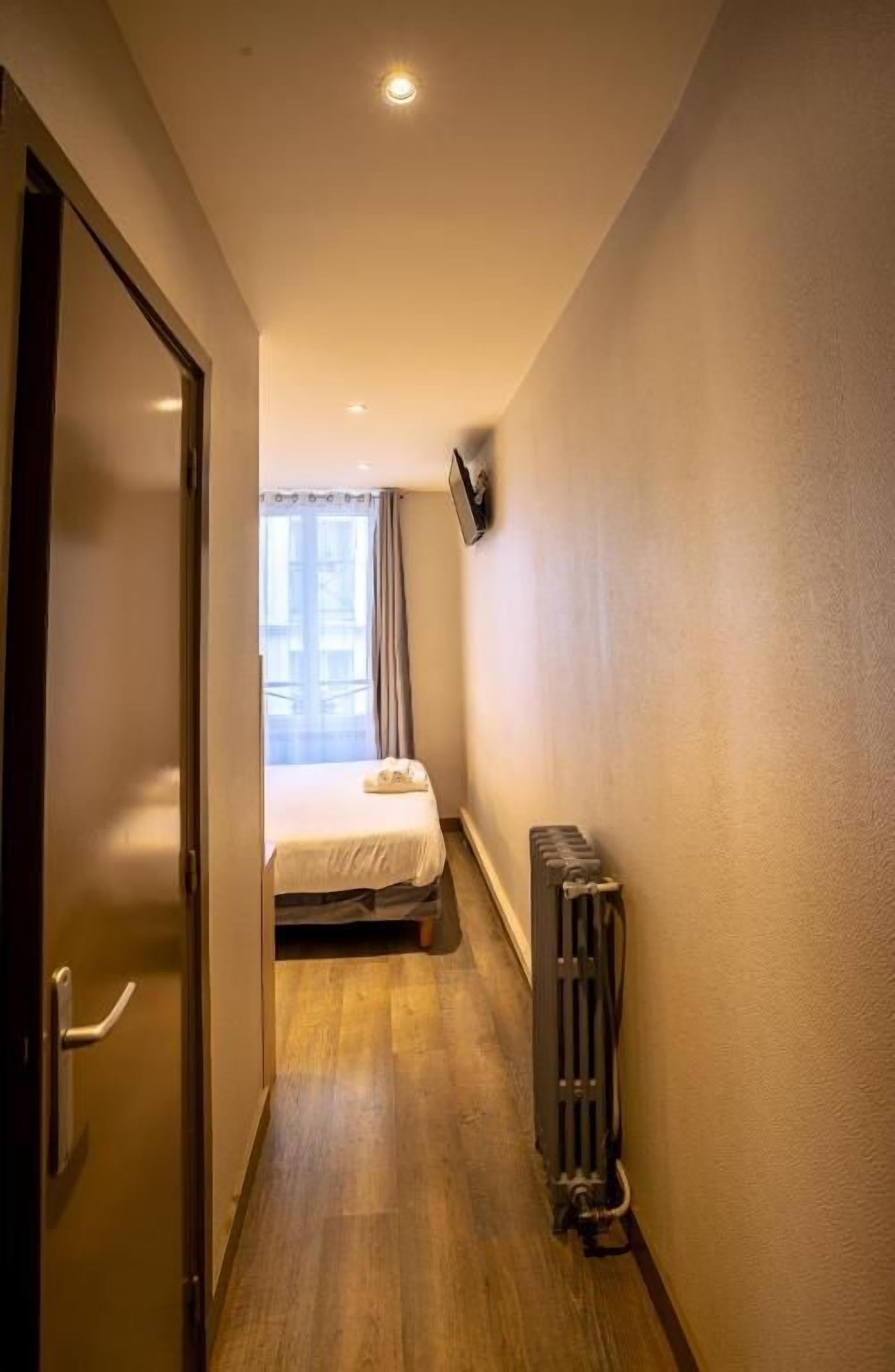 Een smalle gang die leidt naar een bed met een raam, een radiator en een deur aan de linkerkant.