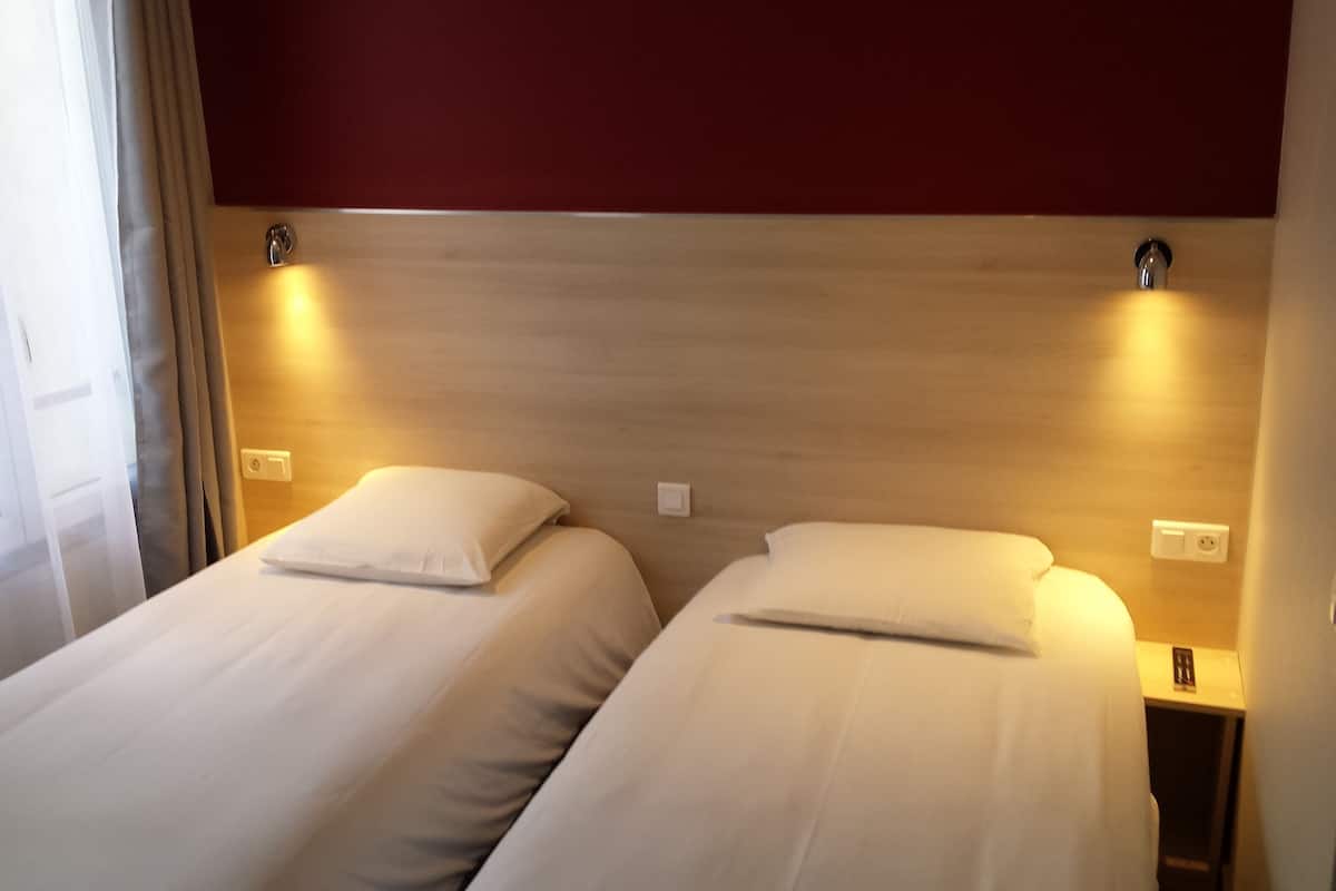 A hotel room with two single beds, wooden headboards, bedside lamps, and a window with curtains.
