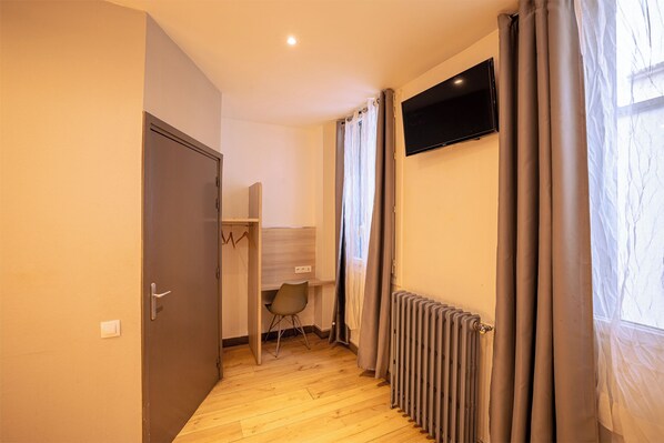A room with a wooden floor, a desk with a chair, a TV mounted on the wall, and a radiator. - Hôtel Aix Europe (Paris)