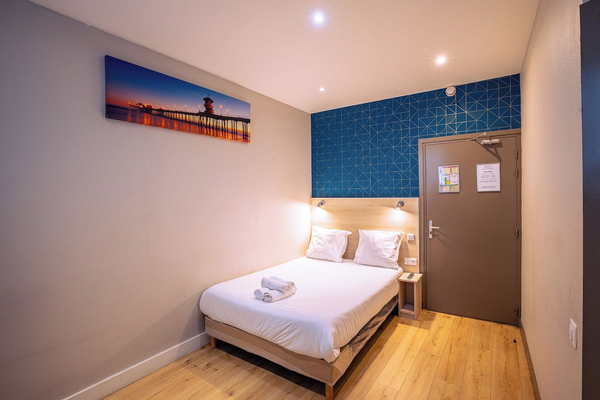A hotel room with a bed, a painting of a sunset, a blue geometric wall, and a door with a notice.