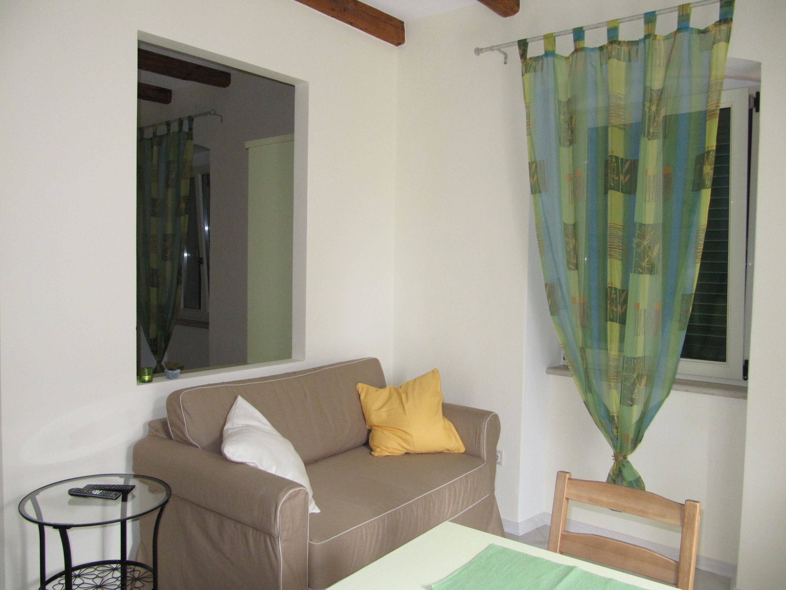 A room with a beige sofa, a small round table with a remote control, a mirror, and a window with a curtain.