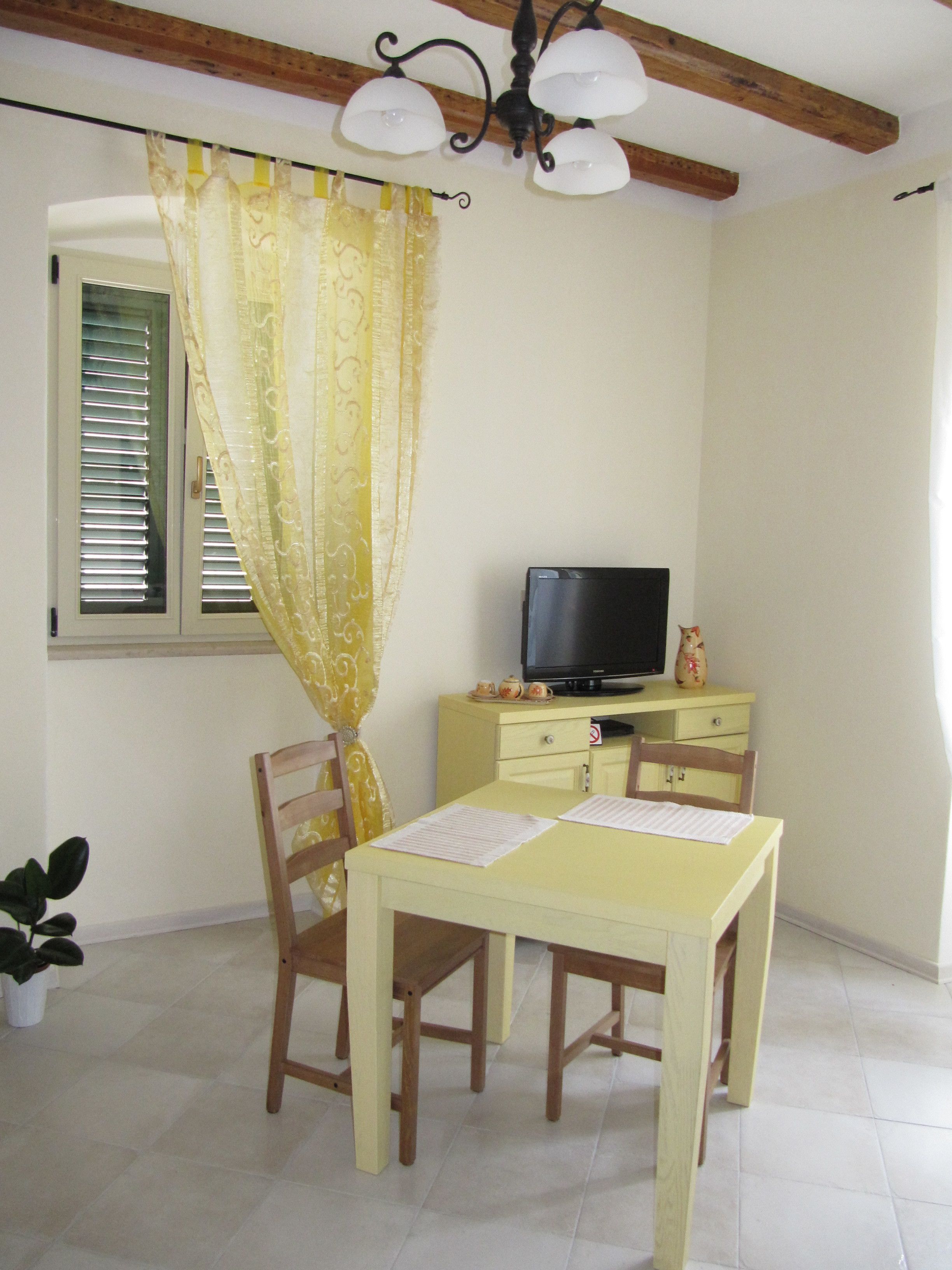 Une pièce comprenant une table à manger, des chaises, un téléviseur posé sur un meuble jaune, une fenêtre avec des stores et un rideau.