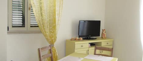A room with a dining table, chairs, a television on a yellow cabinet, a window with blinds, and a curtain.