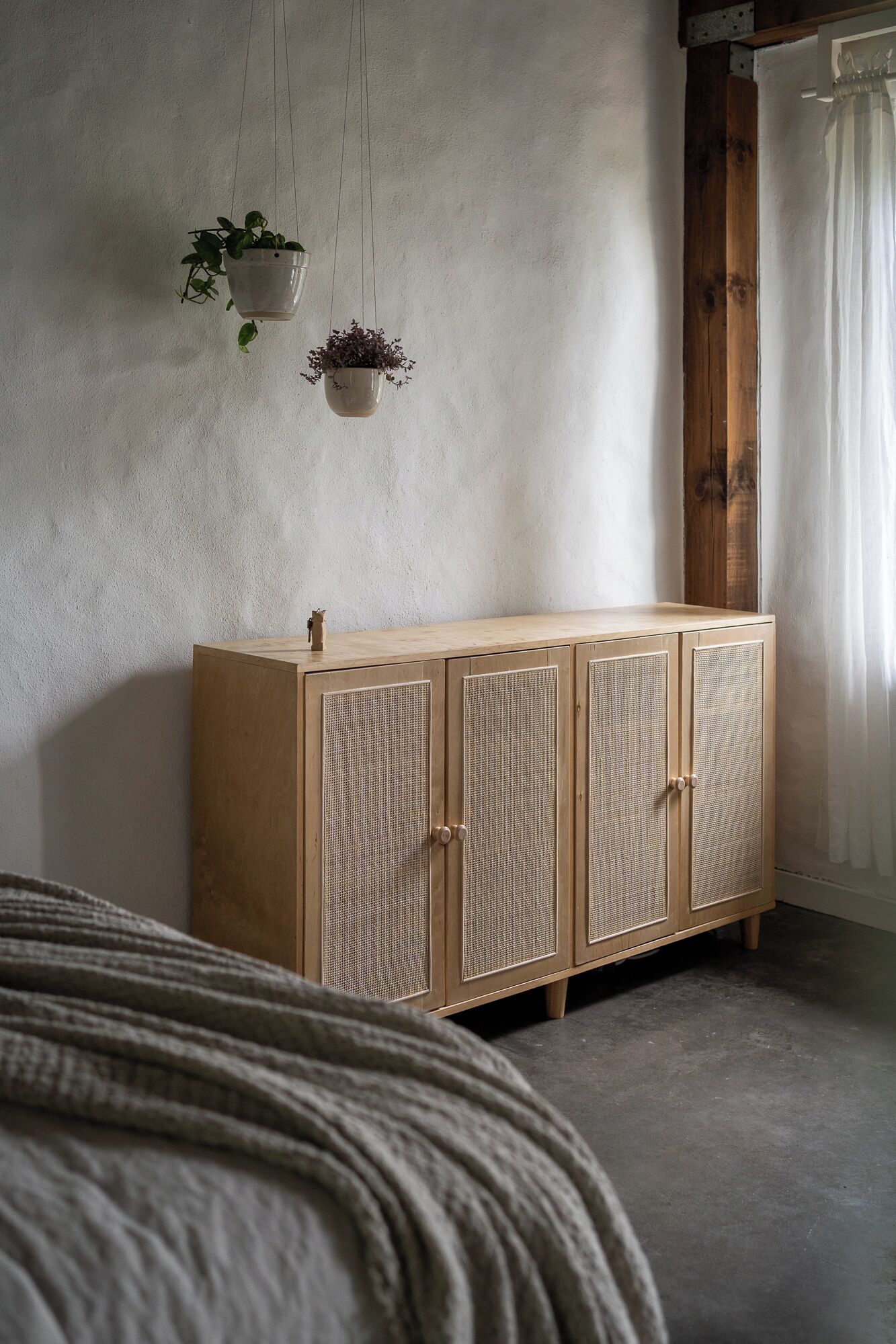 A bedroom with a wooden wardrobe, a hanging plant, and a bed with a grey blanket.