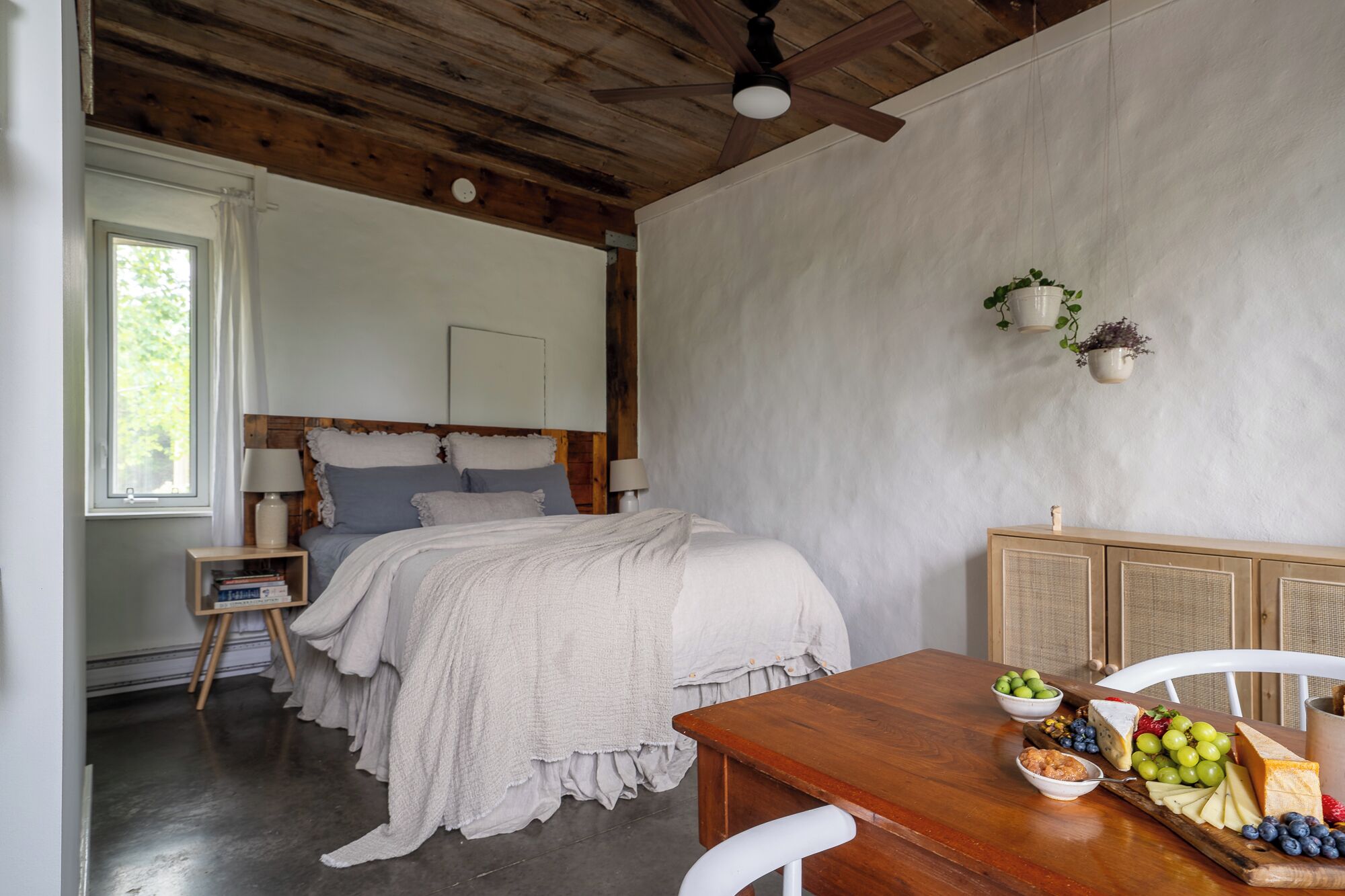 Un dormitorio con techo de madera, una cama con manta blanca, una mesita de noche con lámpara y un aparador de madera con una bandeja de frutas.