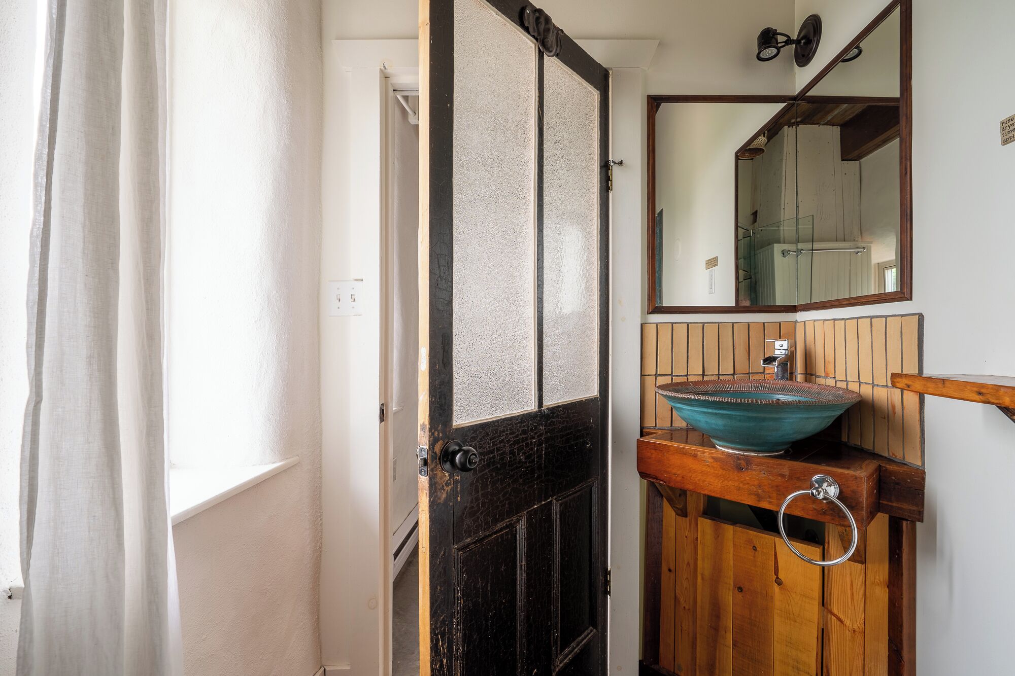 Un baño con un tocador de madera, un lavabo de cerámica azul, un espejo sobre el lavabo y una puerta negra con un panel de vidrio.