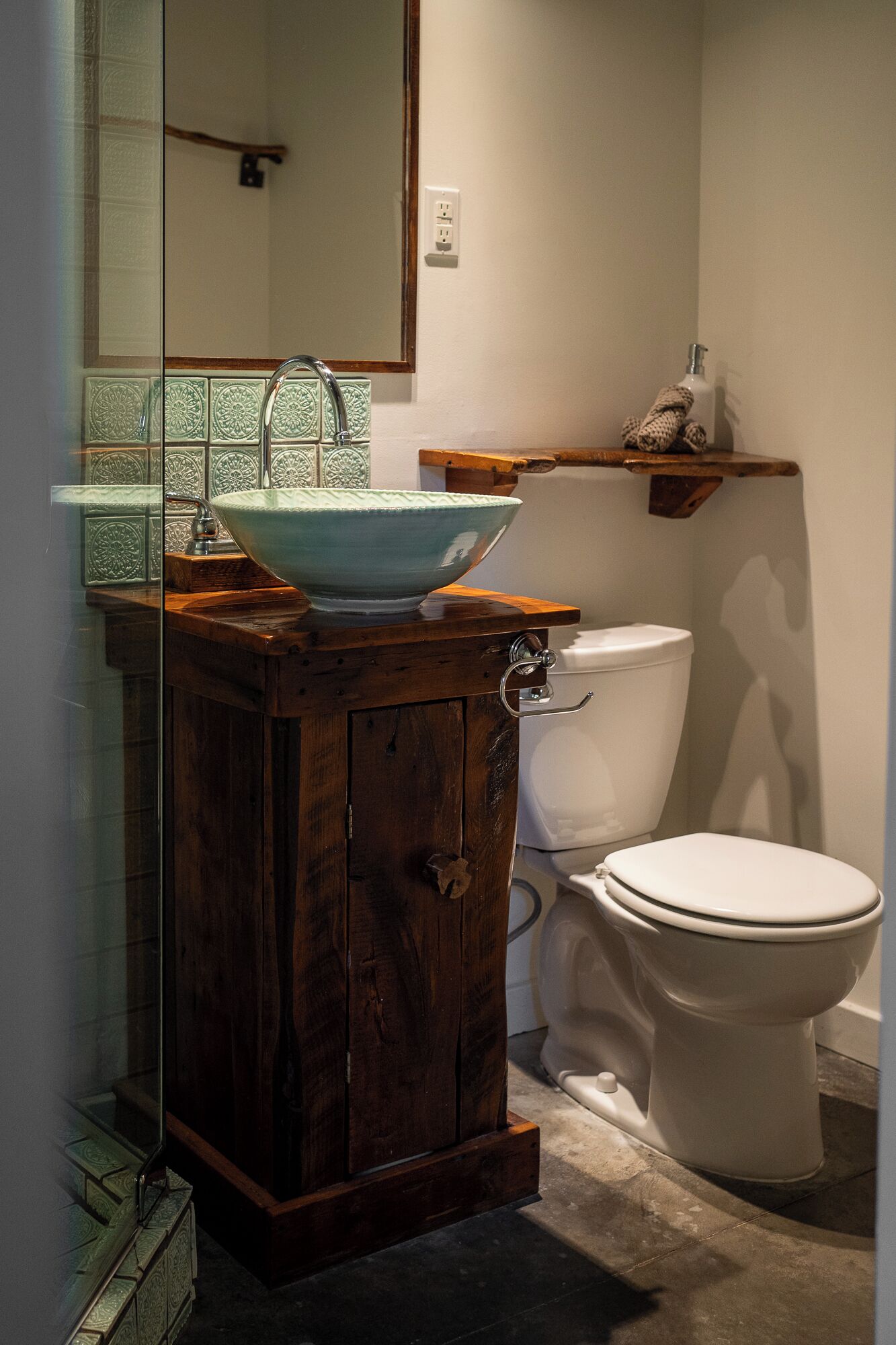 A bathroom with a wooden vanity, a bowl sink, a toilet, and a shelf with toiletries.
