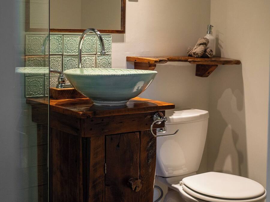 A bathroom with a wooden vanity, a bowl sink, a toilet, and a shelf with toiletries.