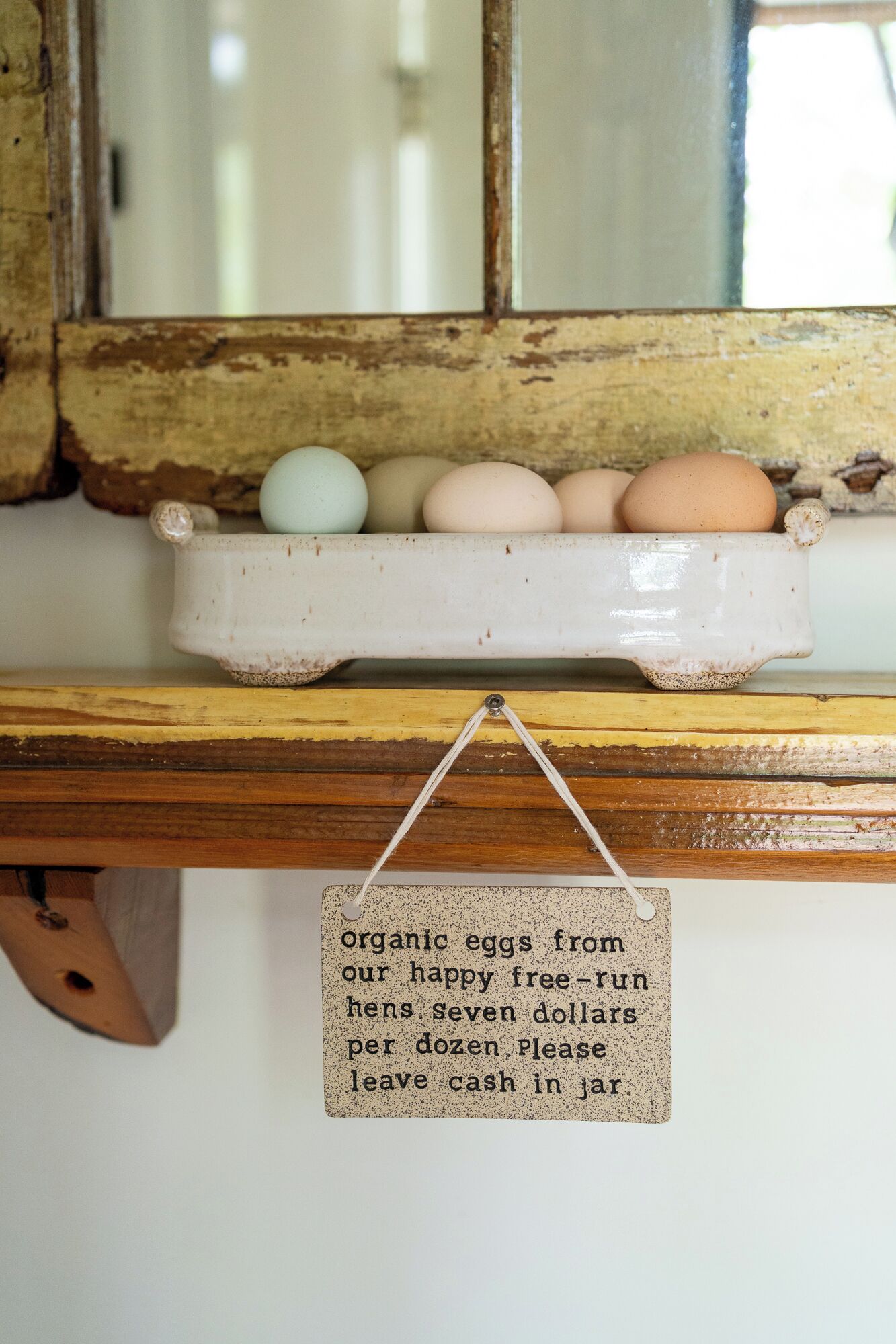 A rustic shelf with a ceramic egg tray holding several eggs, a wooden spoon, and a sign hanging below.