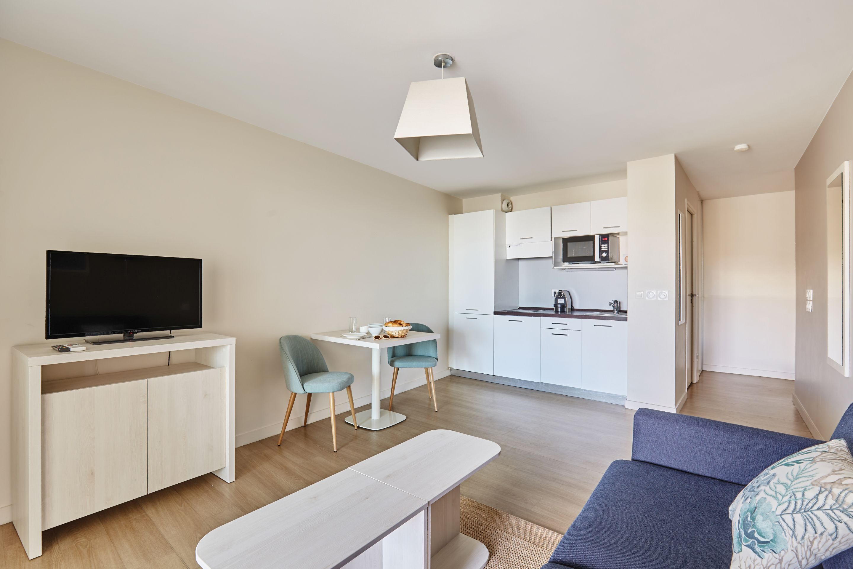 Una sala moderna con televisor de pantalla plana, una mesa de comedor, dos sillas, un sofá y una cocina al fondo.