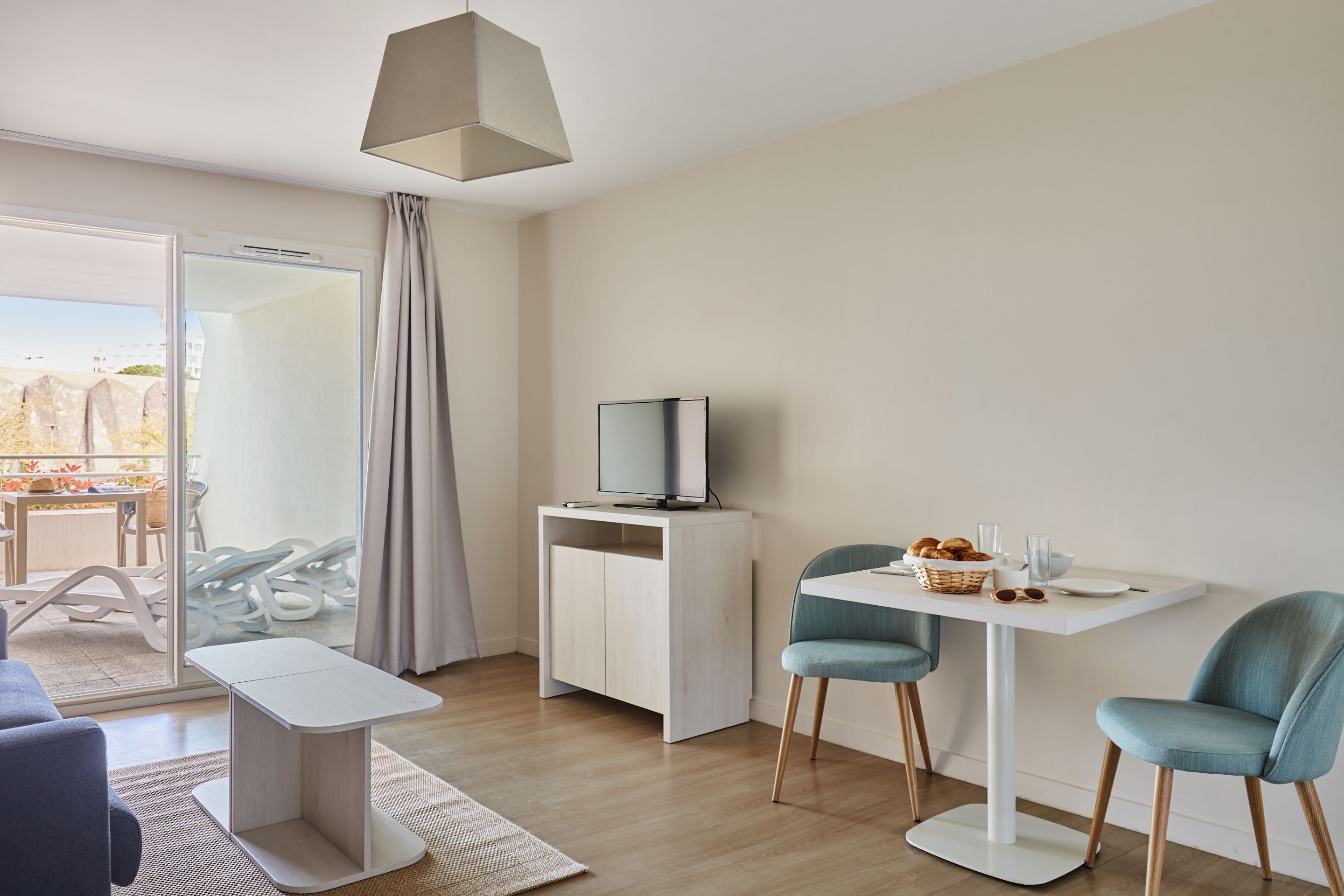 Moderni hotellihuone, jossa on parveke, kaapissa televisio, ruokapöytä tuoleineen ja pieni sohva.