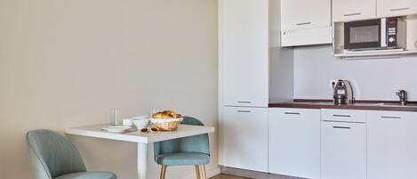 Een compacte keuken met witte kastjes, een magnetron en een koffiezetapparaat. Er staat een kleine eettafel met twee stoelen bij en een mand met brood.