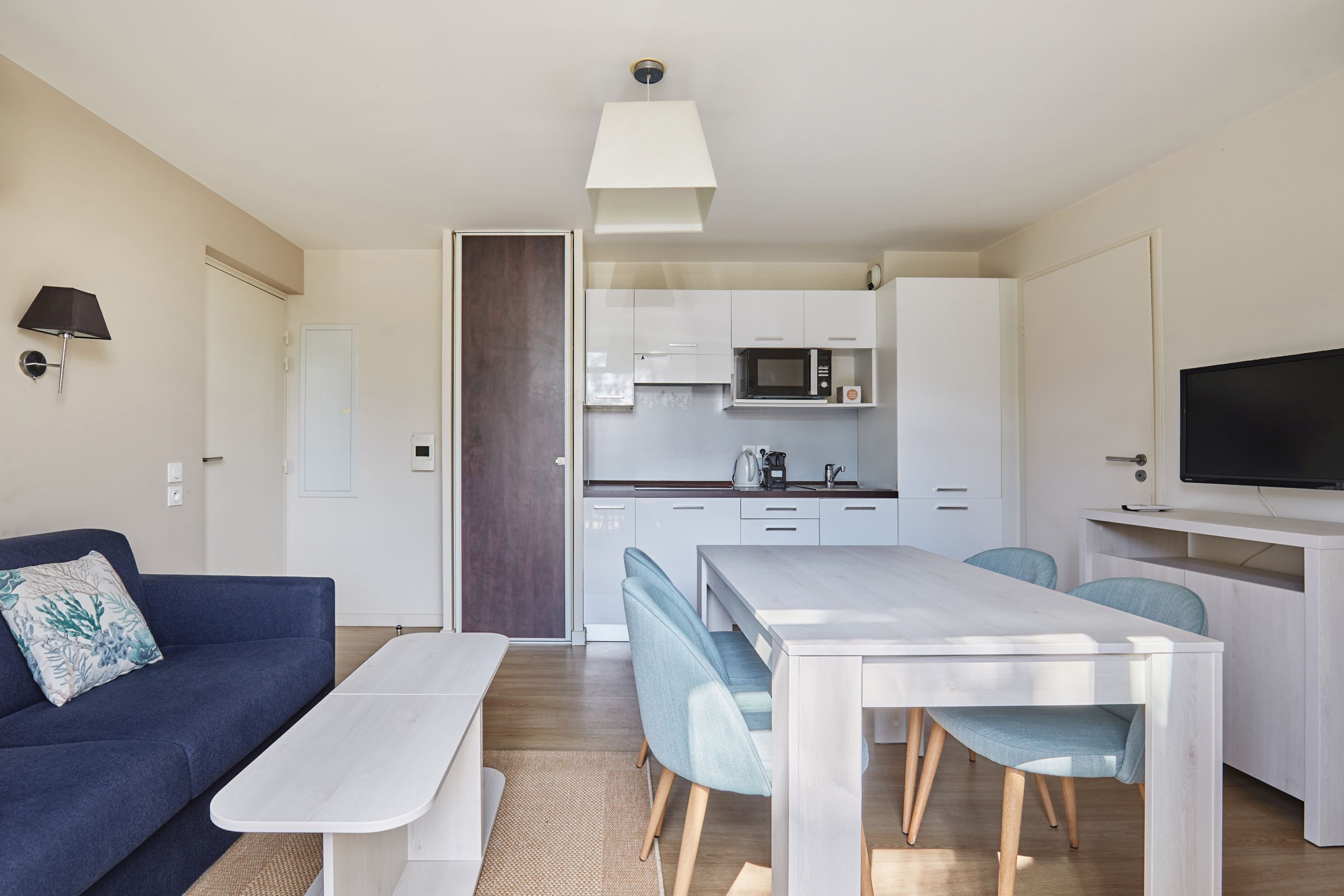 A modern living room with a sofa, a dining table, and chairs. There is a kitchen area with cabinets and a microwave.