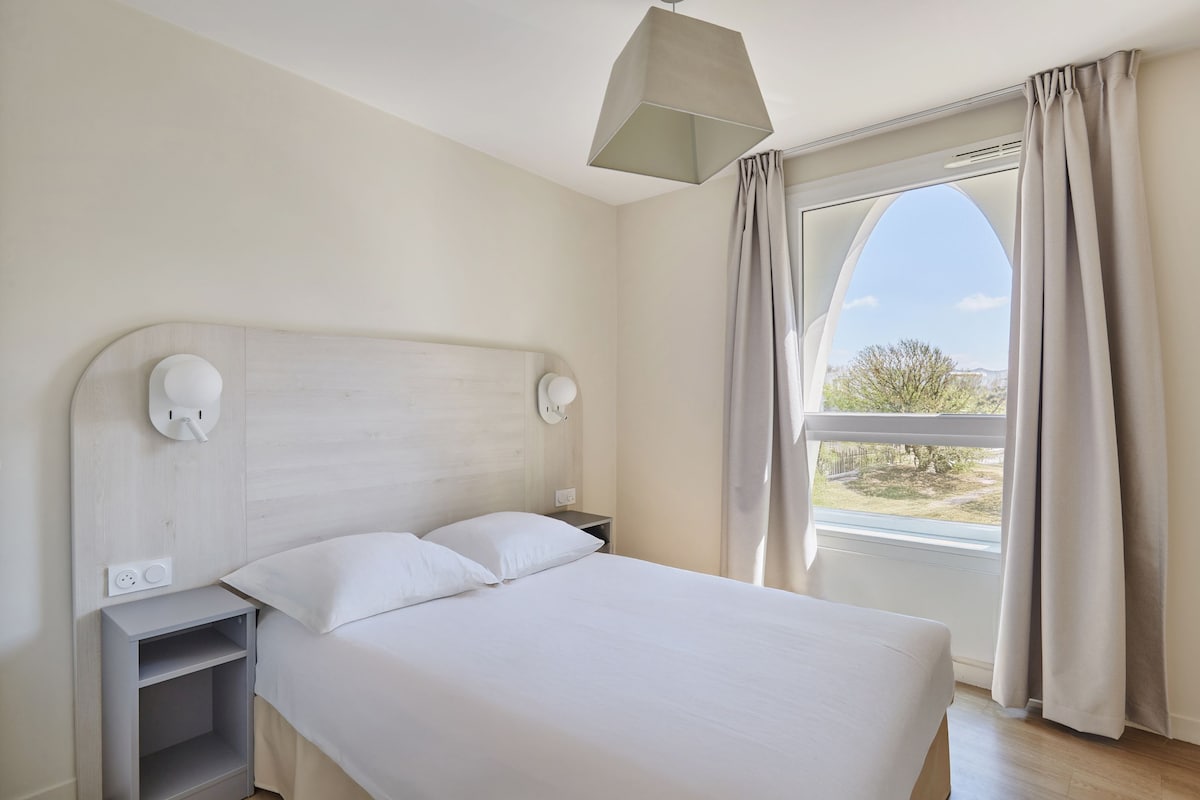 Ein modernes Hotelzimmer mit einem großen Bett, einem Nachttisch, einer Lampe und Blick ins Freie durch ein Fenster.